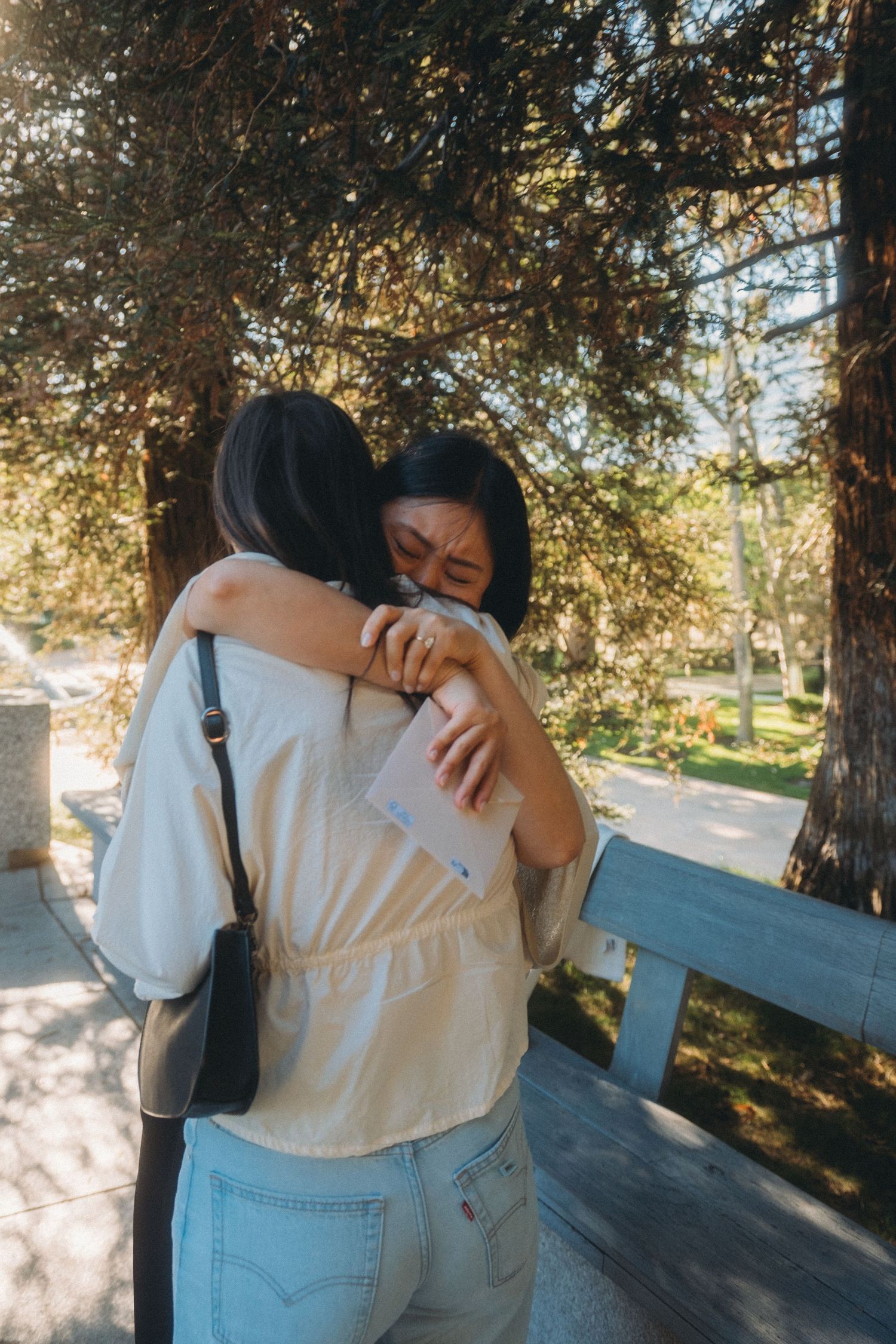 Two young Asian women share an emotional hug outdoors under pine trees in warm sunlight.