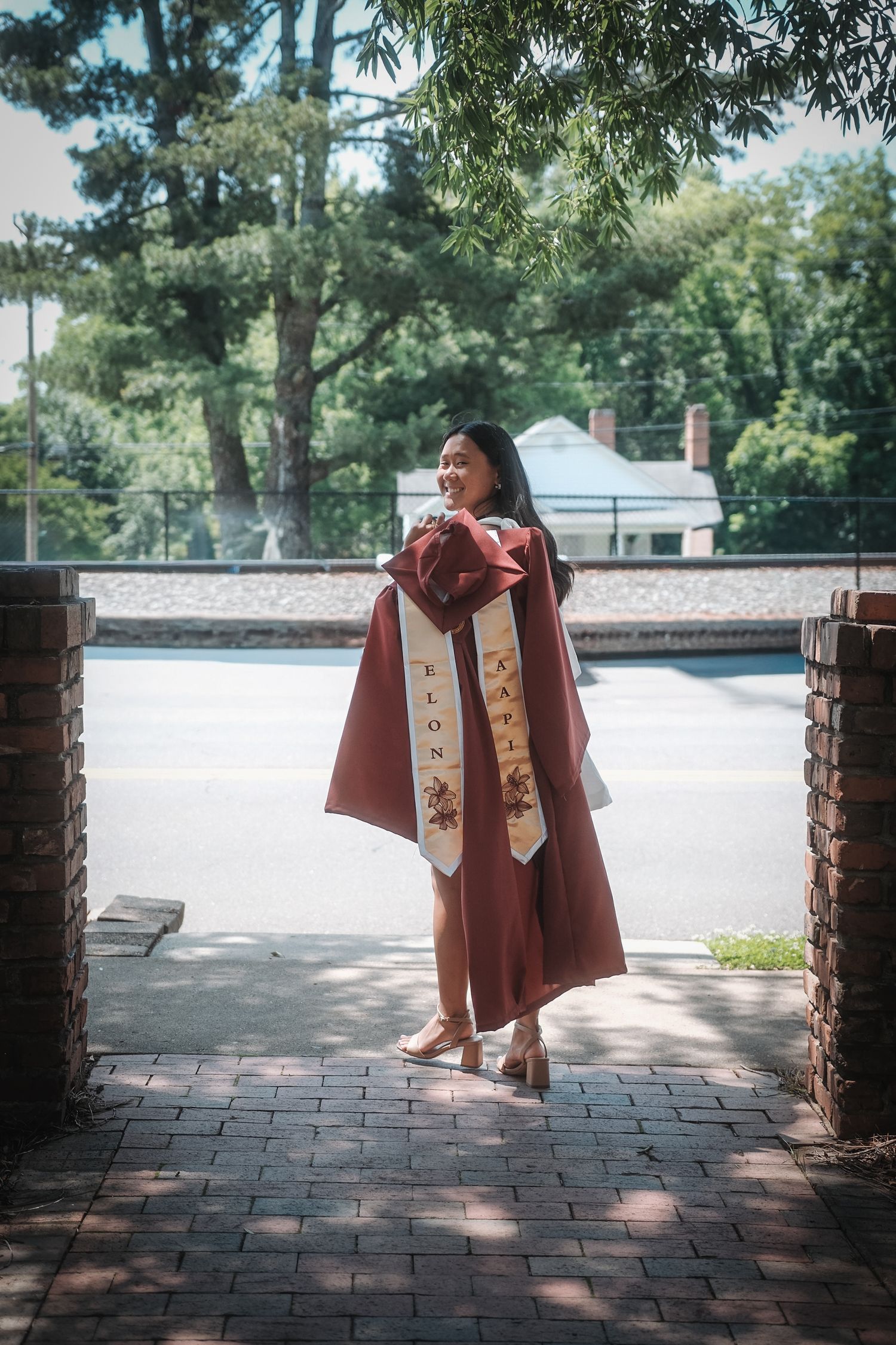 Smiling graduate in maroon Elon University gown poses between brick pillars outdoors.