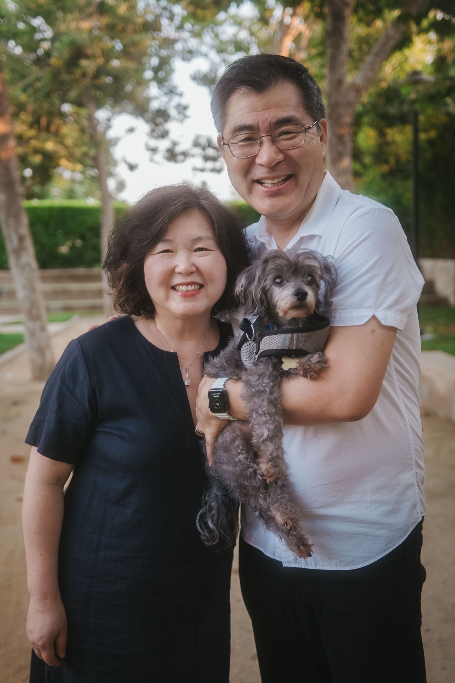 Smiling couple posing outdoors with a small gray poodle mix dog in a park setting.