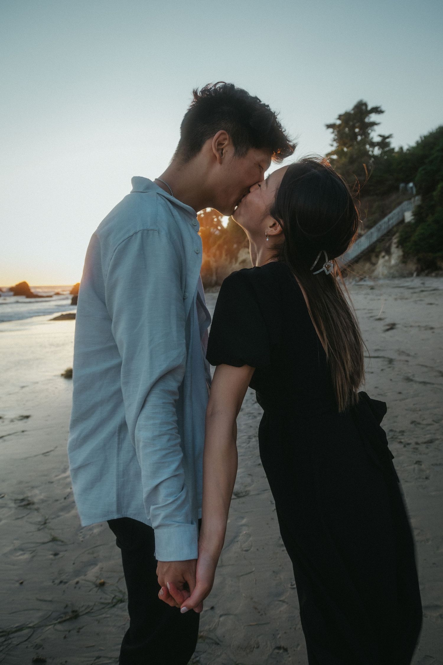 Couple sharing a romantic almost-kiss on a beach at sunset, man in blue shirt and woman in black dress holding hands.