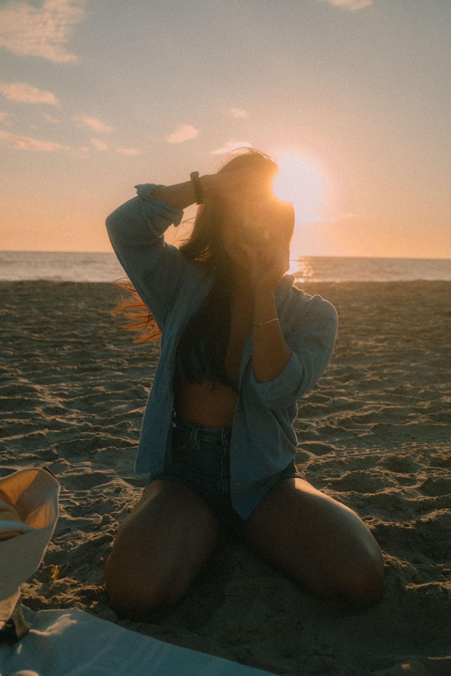 Silhouetted woman kneeling on beach blanket with hands in hair, backlit by stunning golden ocean sunset.