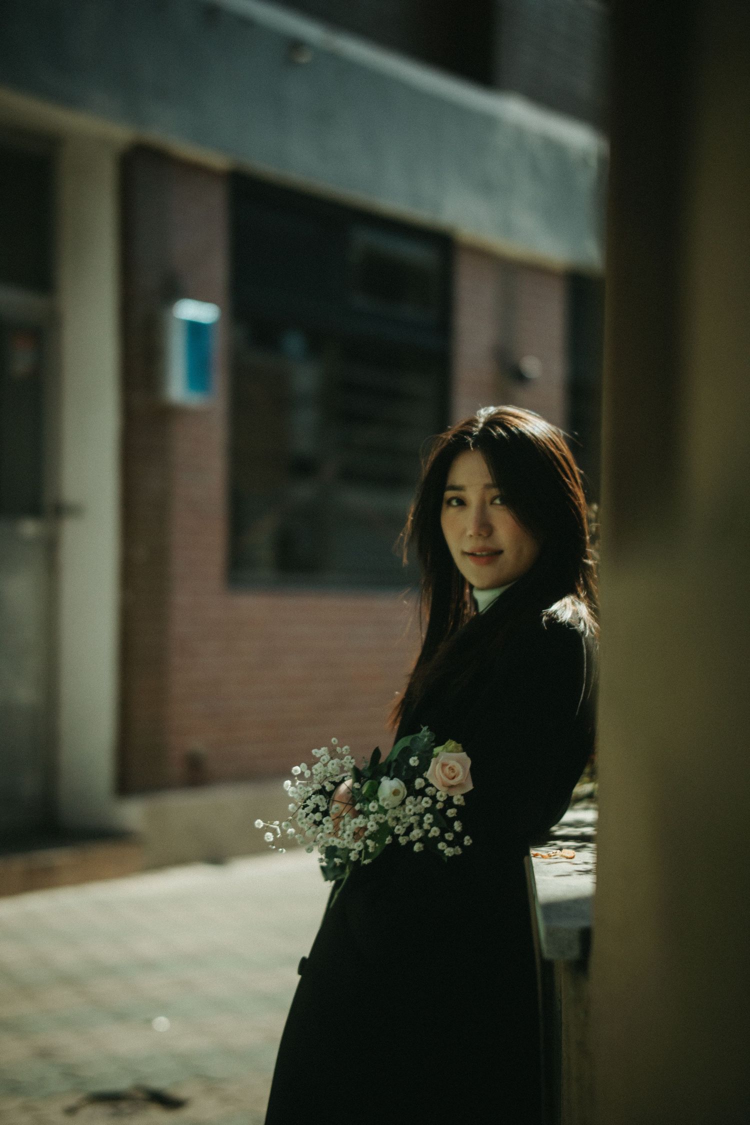 Young Asian woman in black coat holding a rose bouquet, posing in a dimly lit urban alley.