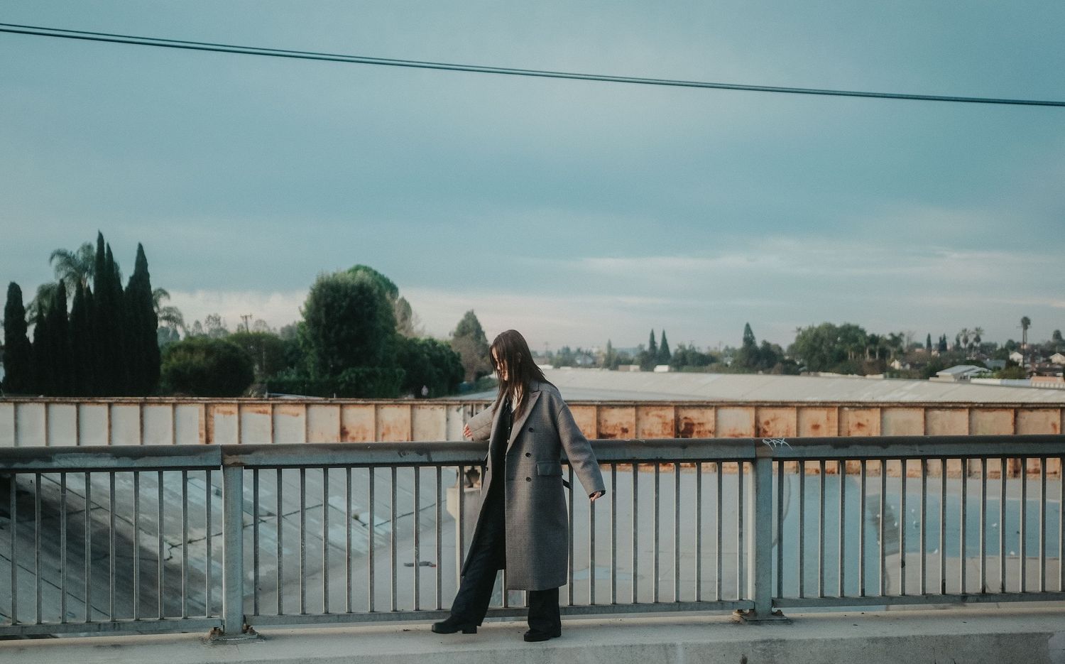 Woman in long gray coat standing on a bridge railing overlooking a scenic waterway on a cloudy day.