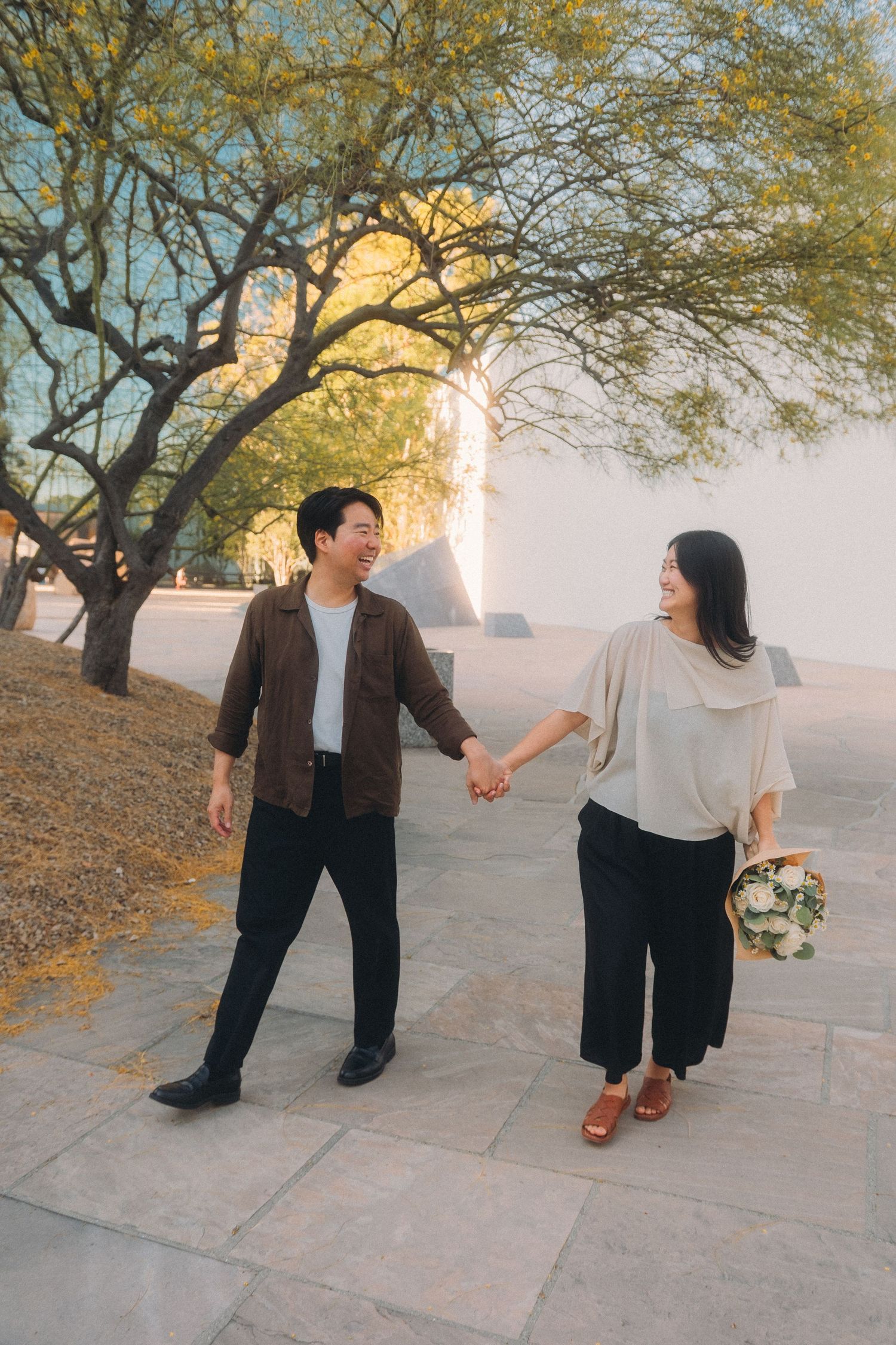 Joyful Asian couple holding hands and smiling at each other on sunlit stone walkway under green trees.