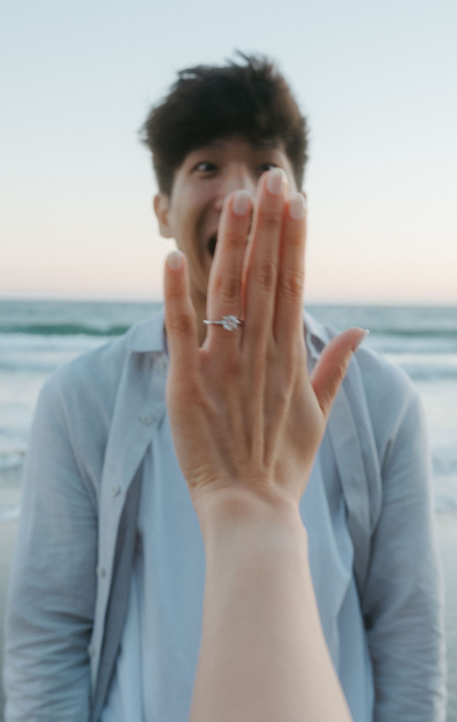 Woman showing off diamond engagement ring to camera with excited man in background at beach.