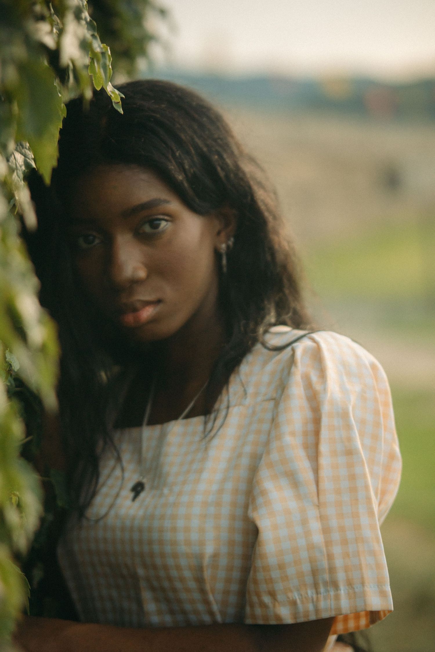 A young Black woman with long wavy hair peers through green leaves wearing a light gingham blouse.