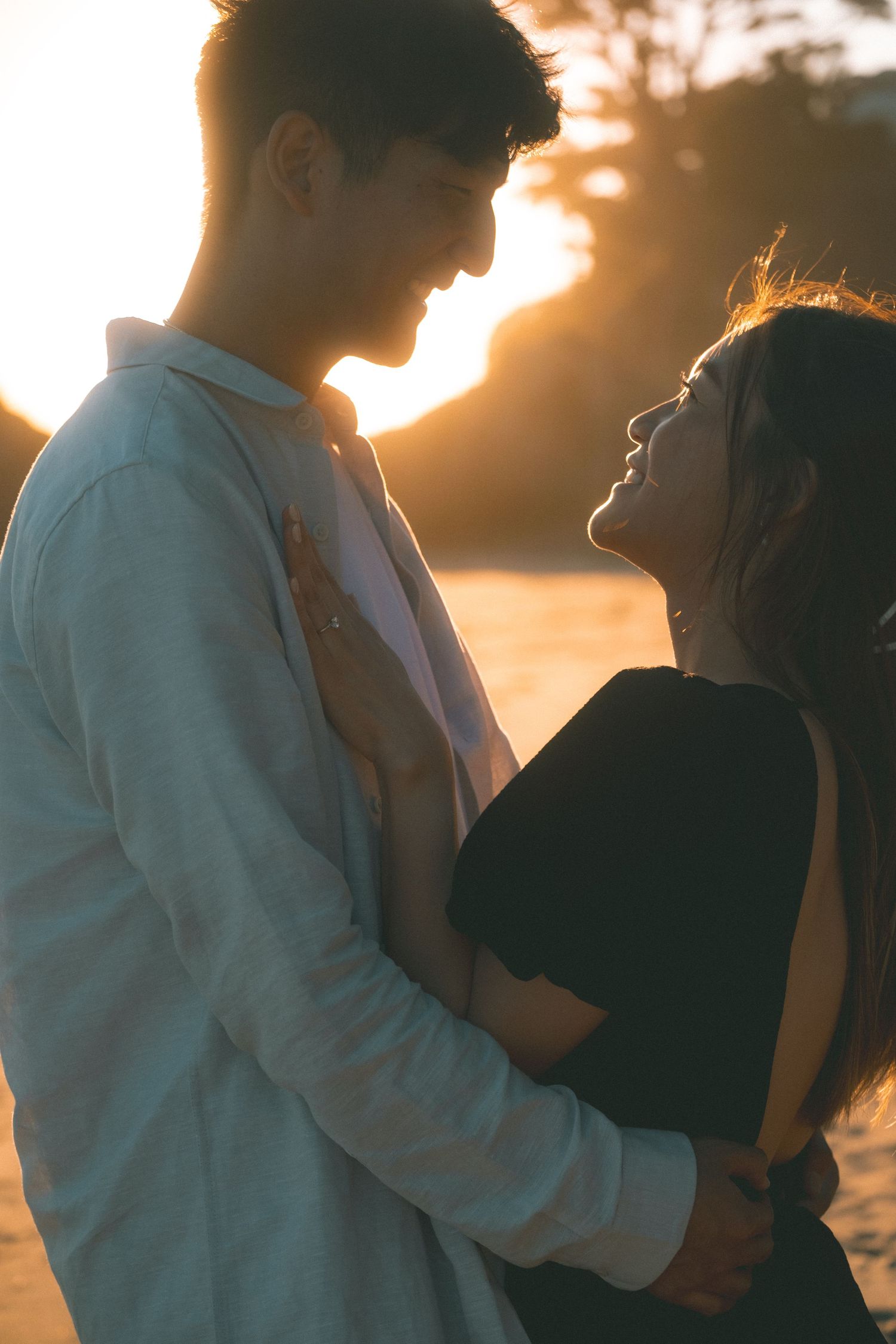 Romantic couple gazing at each other while embracing on a beach during a warm golden sunset.