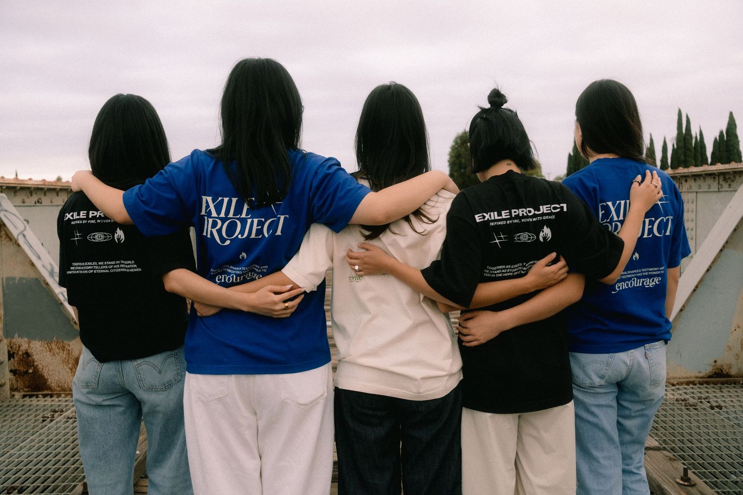 Five young women with arms around each other wearing Exile Project blue and black t-shirts, viewed from behind outdoors.
