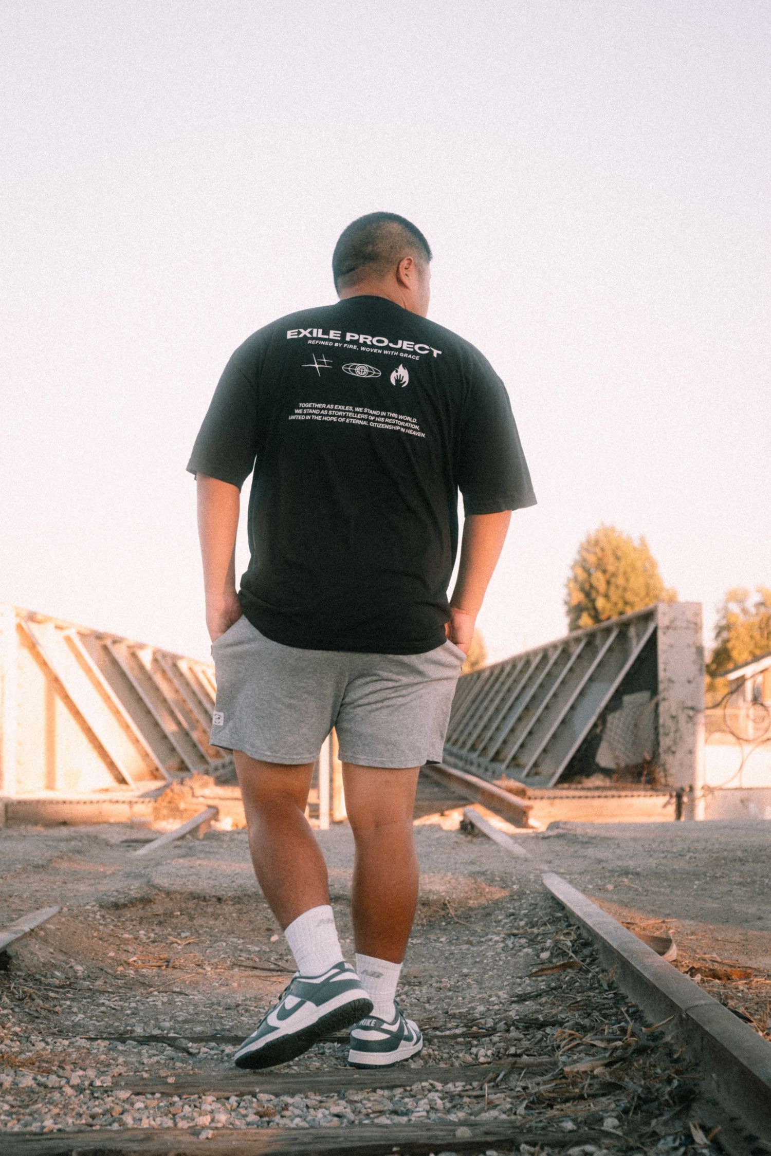 Man wearing black Exile Project t-shirt and grey shorts standing on railroad tracks outdoors.