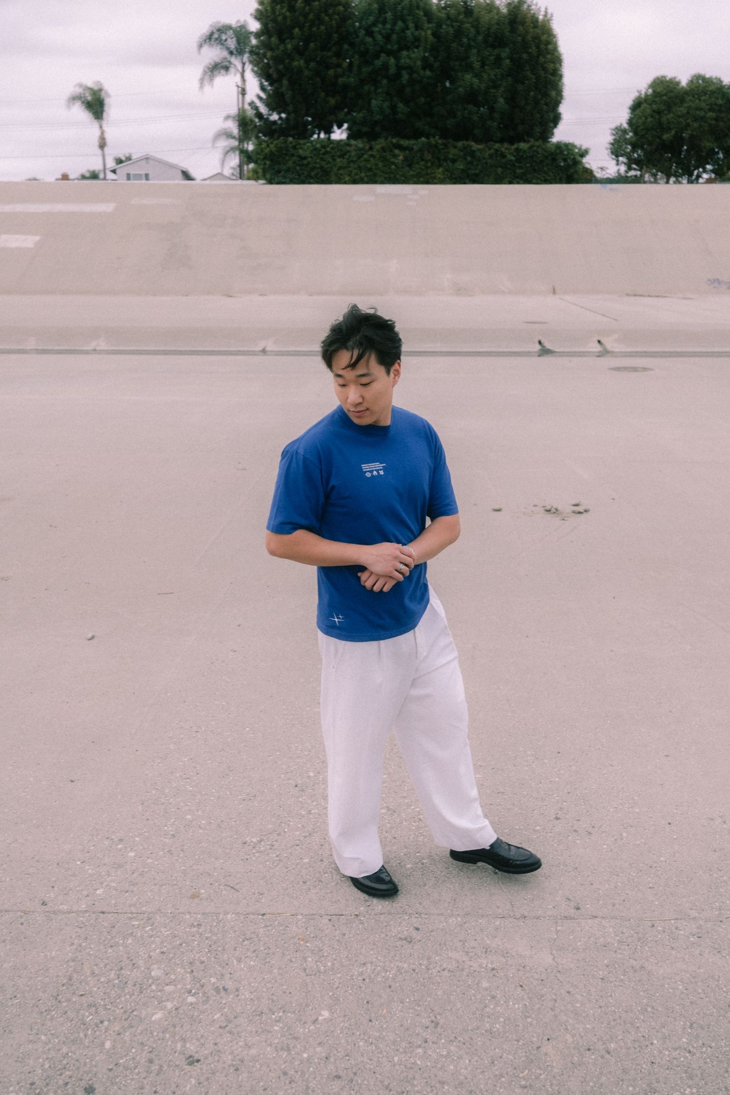 Young man in blue graphic tee and white wide-leg pants looking down on an outdoor concrete surface.