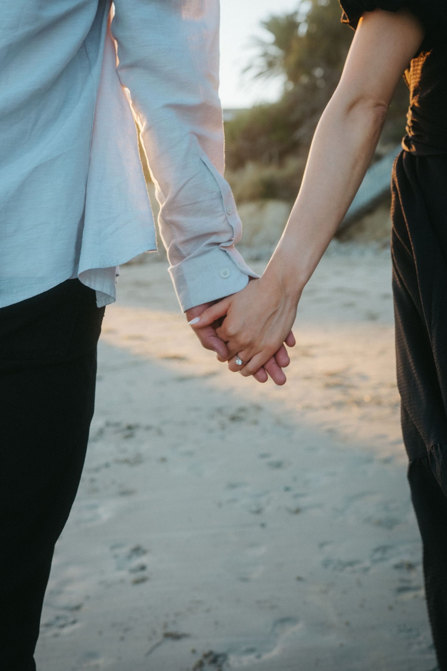 Close-up of an engaged couple holding hands on a sandy beach, showing a diamond engagement ring.
