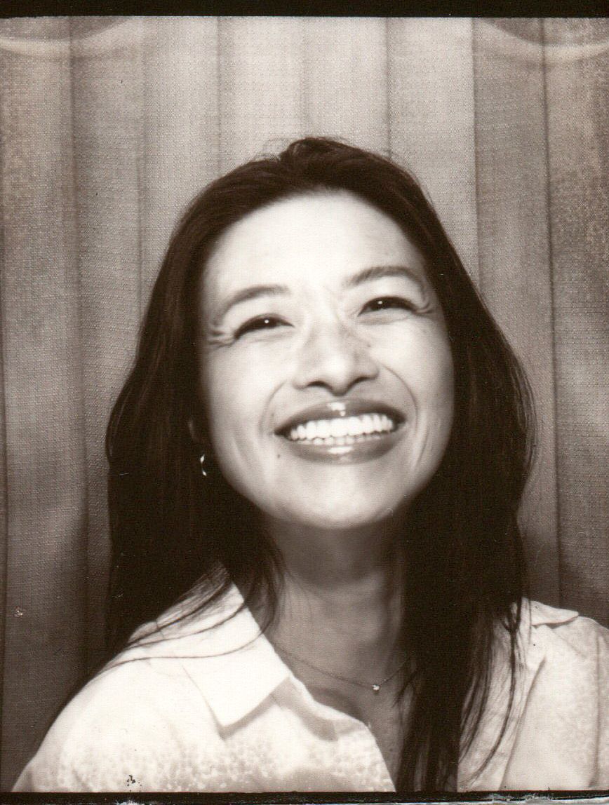 Smiling woman with long dark hair in a vintage black and white photo booth portrait, wearing a white shirt.