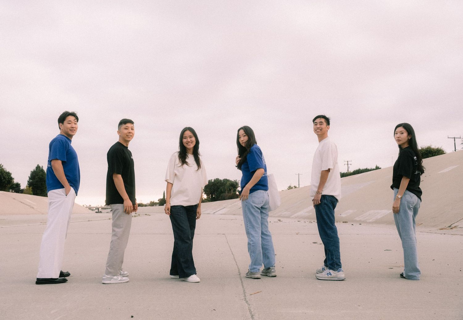 Group of six young adults in casual streetwear posing together at an outdoor skate park under cloudy skies.