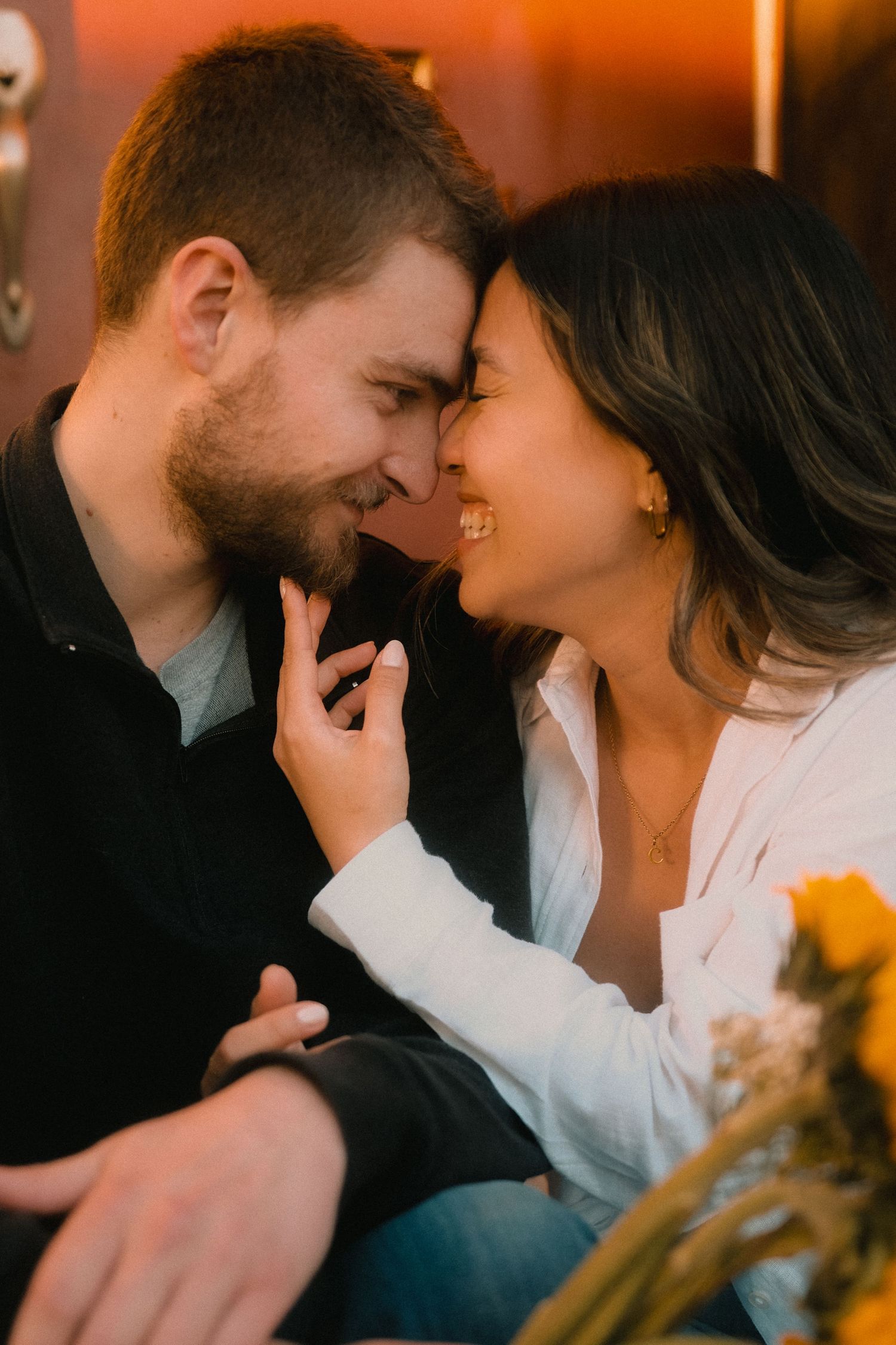 A couple shares a romantic moment, smiling and touching foreheads in warm golden light with flowers nearby.