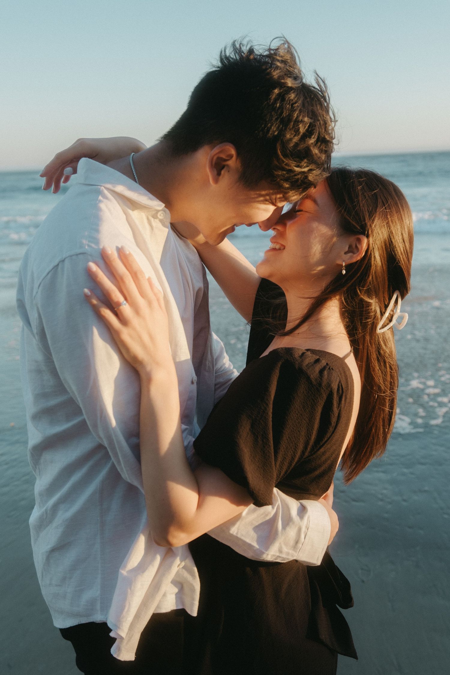 Couple smiling and embracing face-to-face on a sunlit beach, woman wearing an engagement ring.