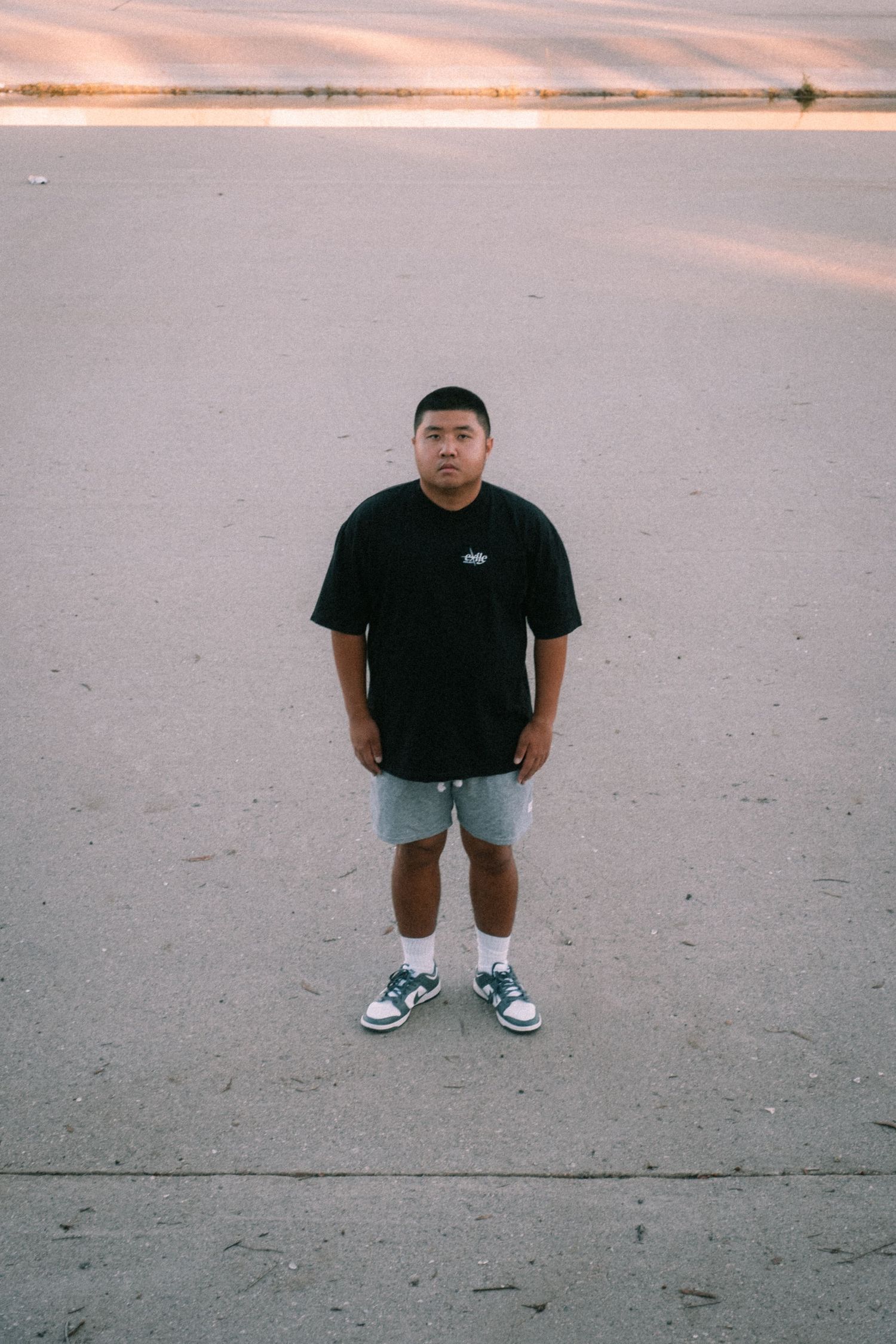 Young man in black oversized tee, grey shorts, and Nike sneakers standing on concrete near a beach at sunset.