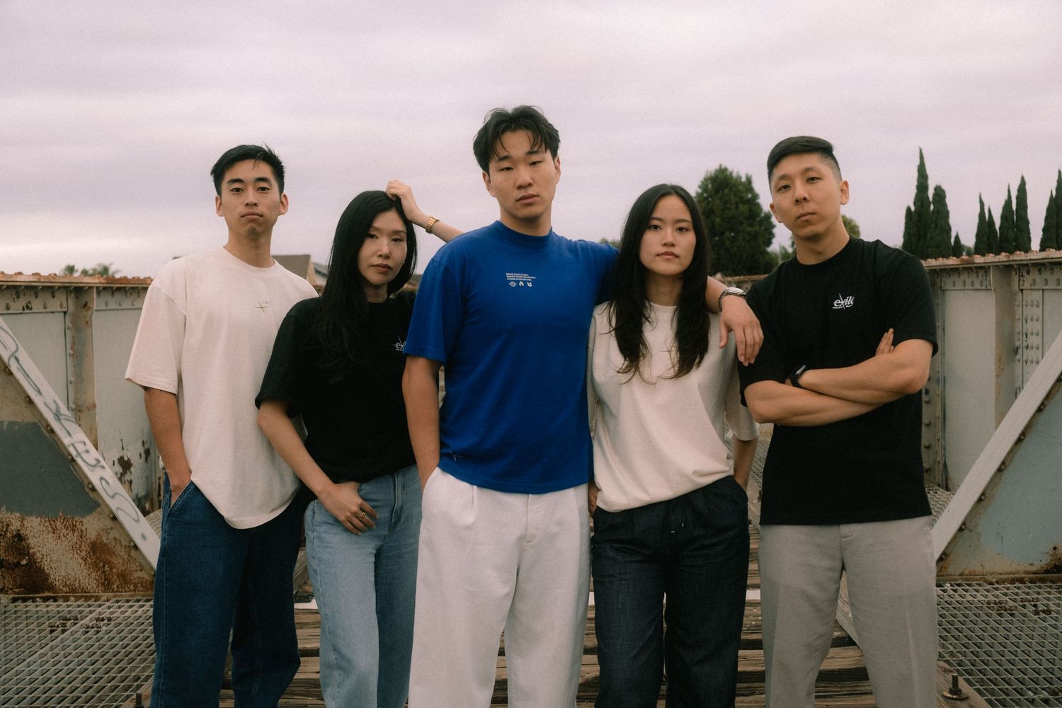 Group of five young adults posing in casual streetwear on a rooftop under a cloudy sky.