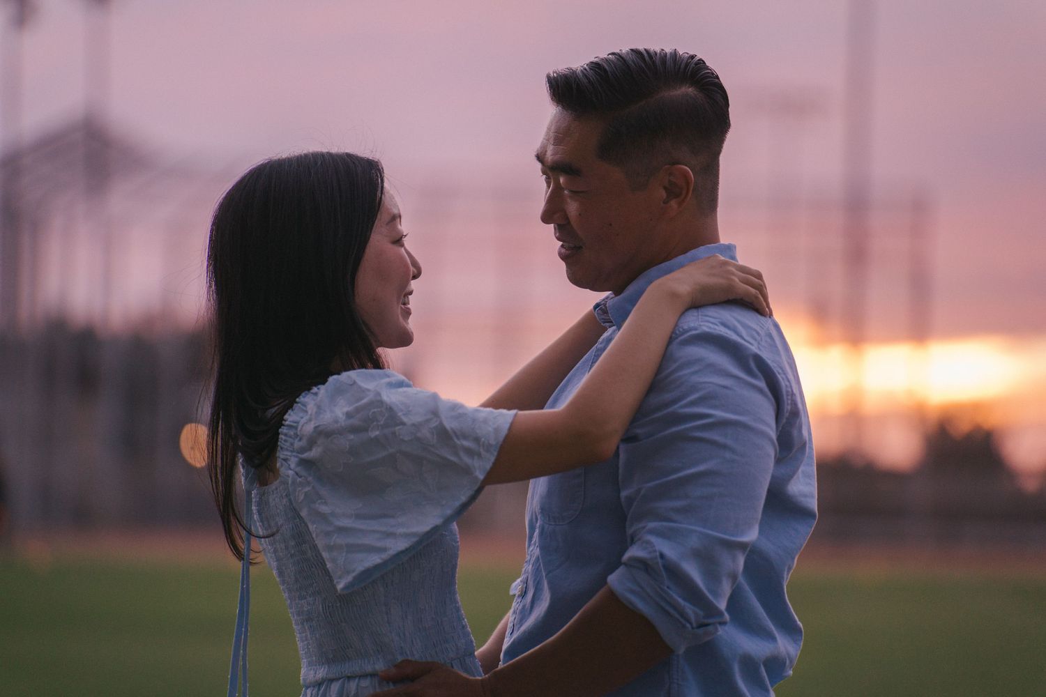 Couple embracing at sunset on a field, sharing a romantic moment with a colorful pink sky in the background.