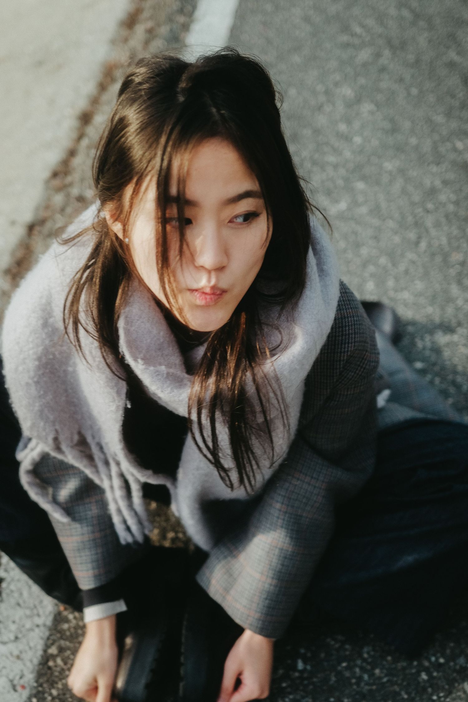 Young Asian woman in gray coat and scarf sitting on pavement, looking up with windswept hair.