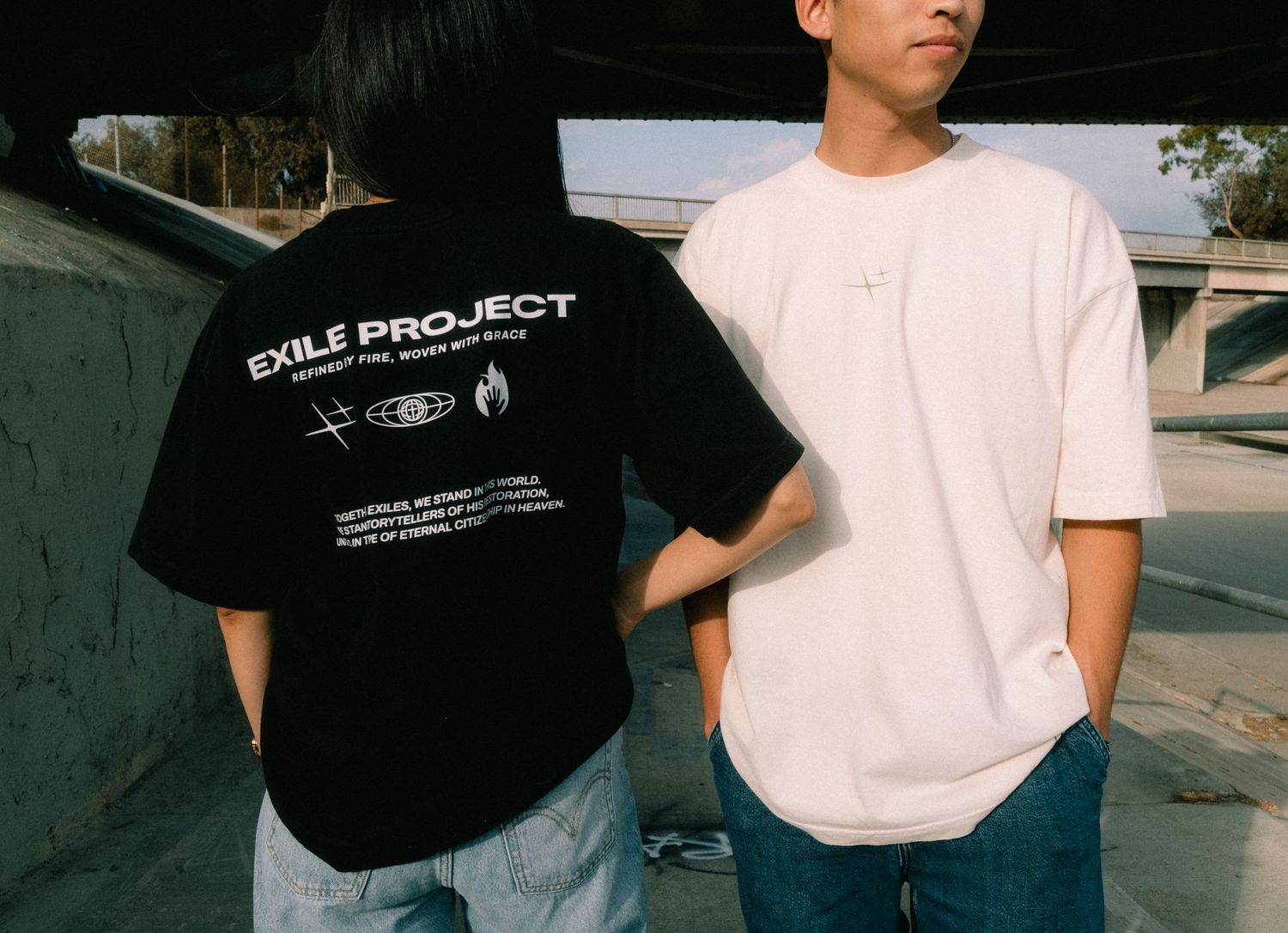Two models wearing Exile Project graphic t-shirts in black and white, posed back-to-back outdoors.