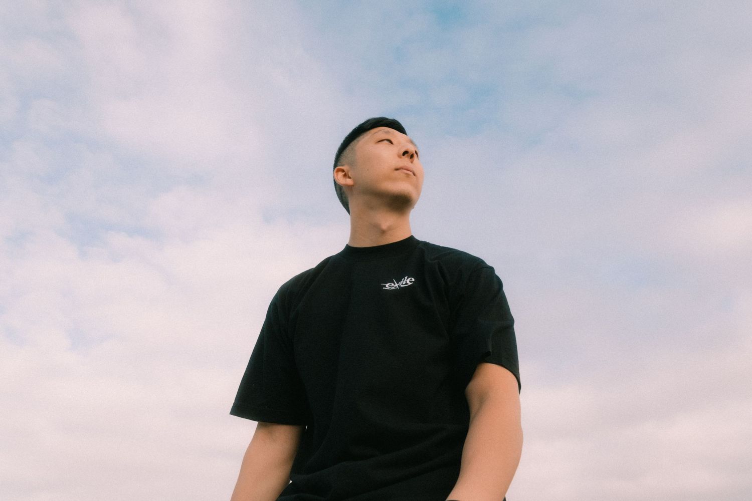 Young man in black t-shirt gazing upward against a cloudy blue sky backdrop.