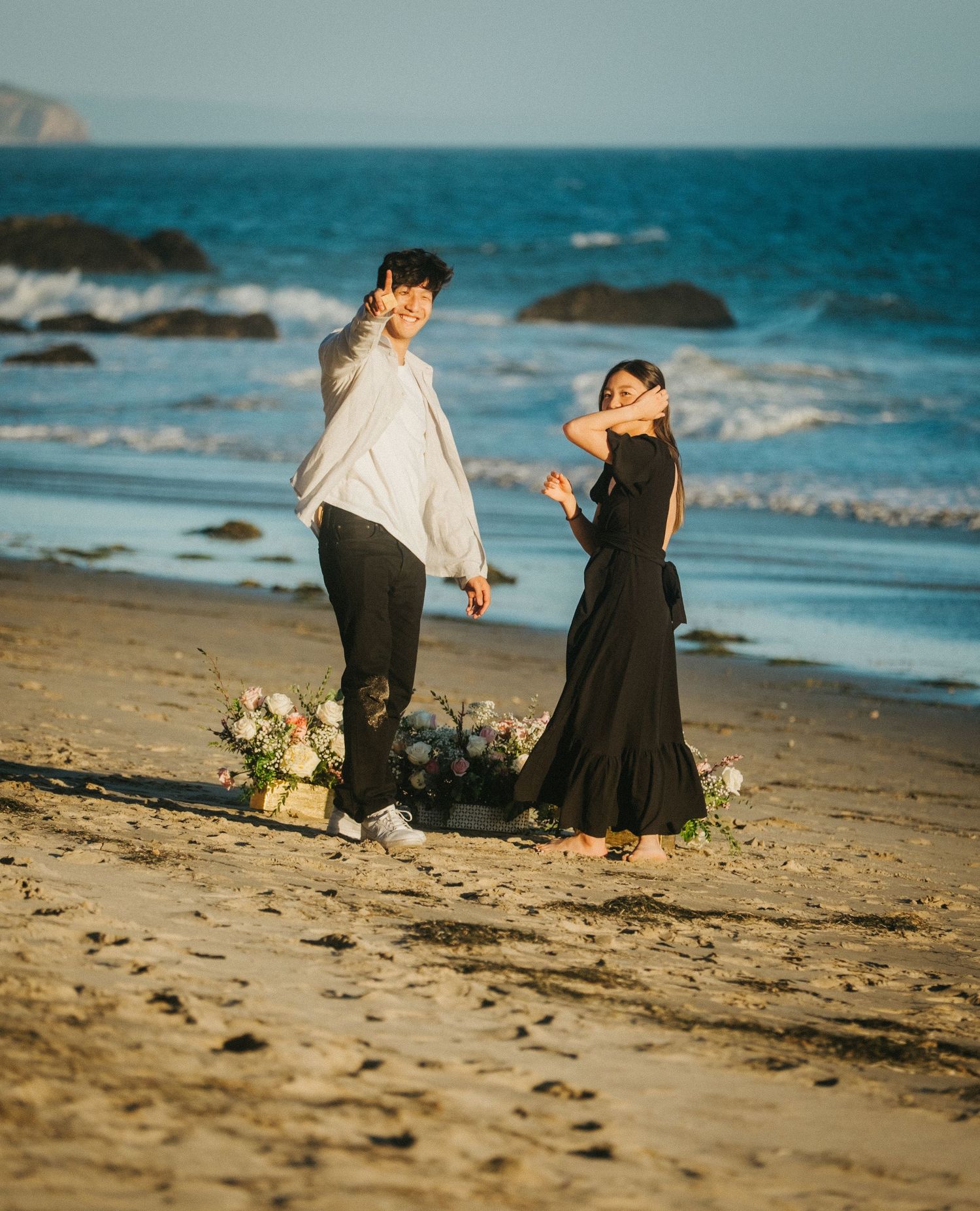 Engaged couple celebrating playfully on a sunny beach with floral decorations and ocean backdrop.