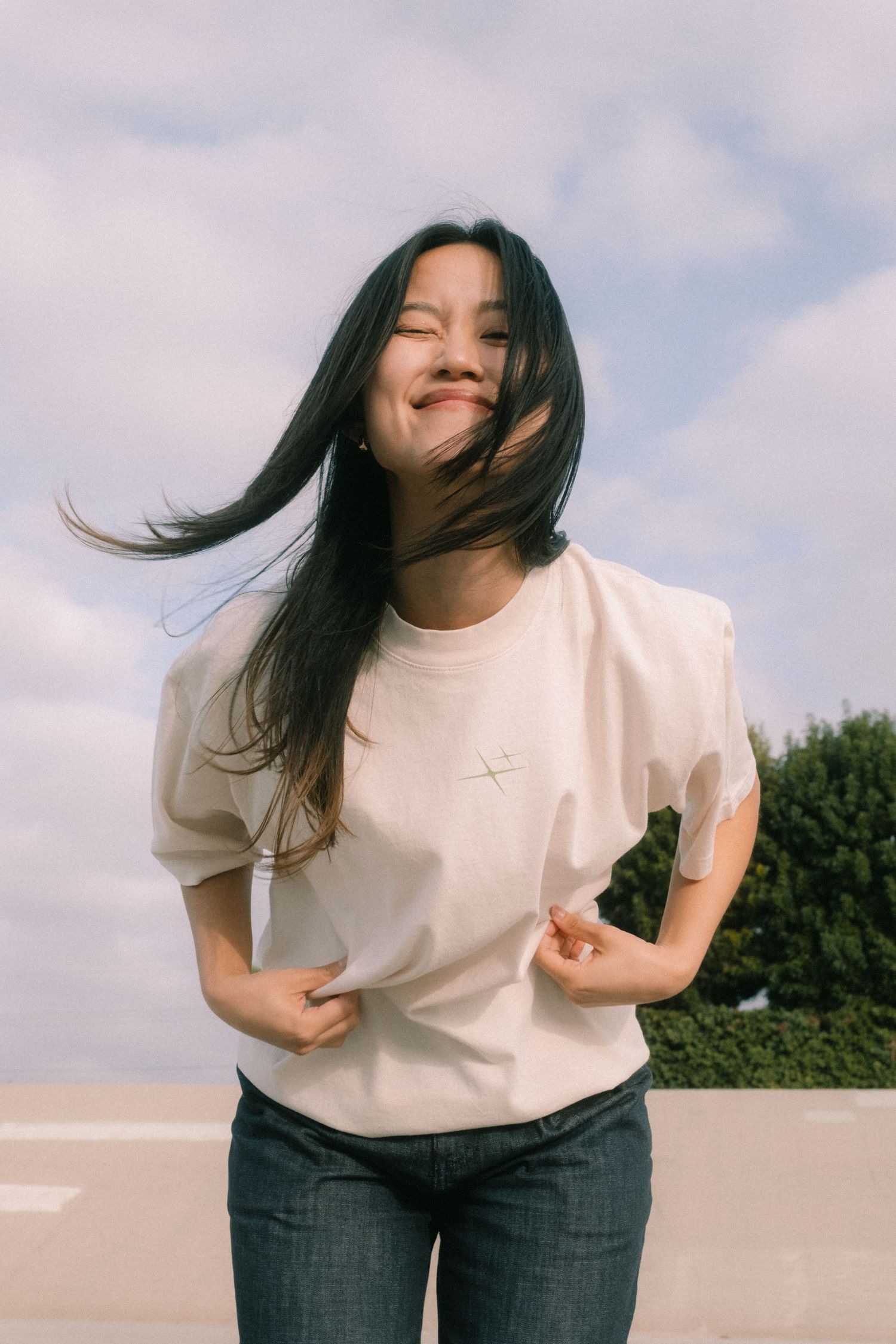 Smiling young woman with wind-blown hair wearing a cream puff-sleeve tee and dark jeans outdoors.