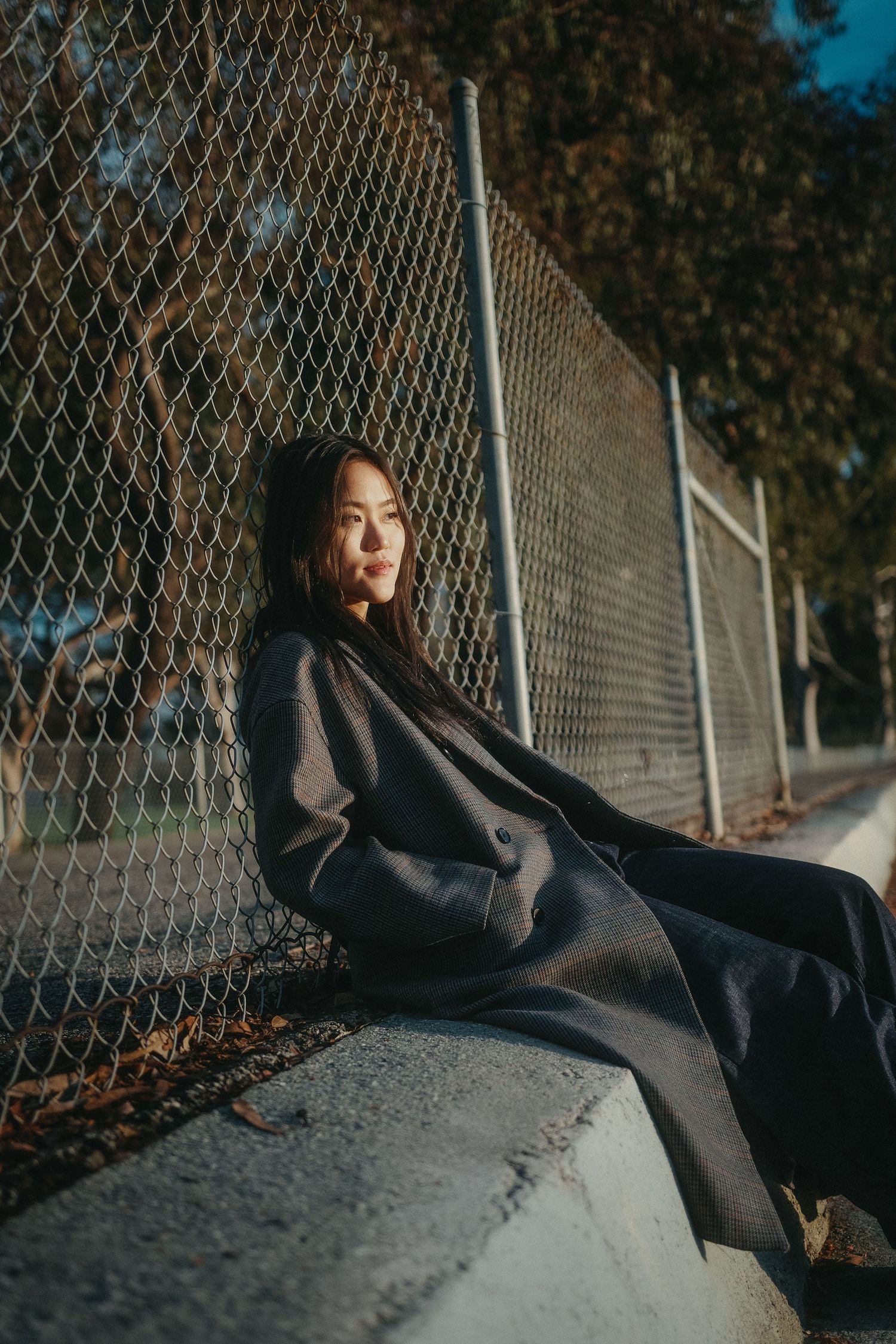 Young Asian woman in a long dark coat sitting against a chain-link fence in warm sunlight.
