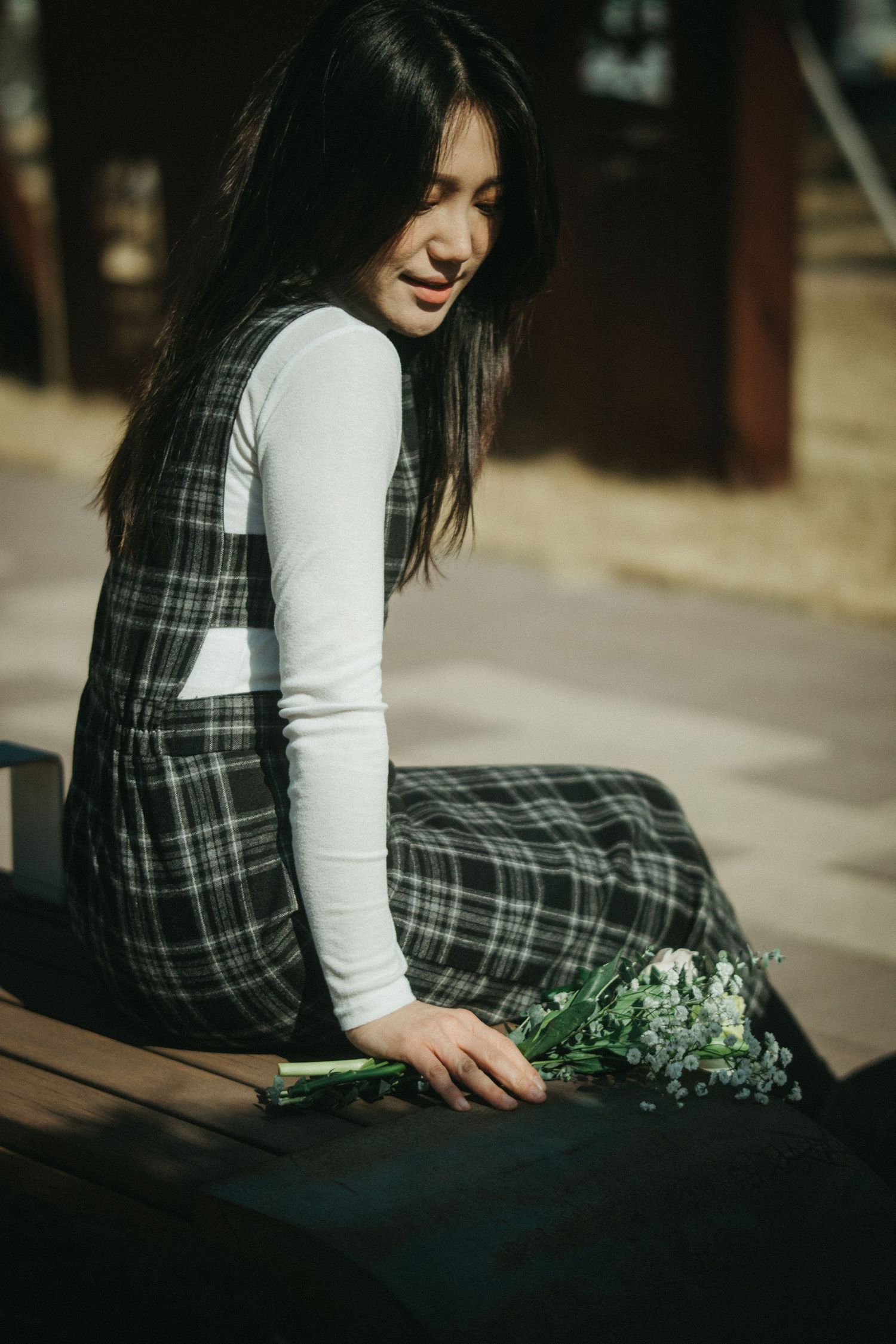 Young Asian woman in plaid dress sitting on a bench, looking down at a bouquet of white flowers.