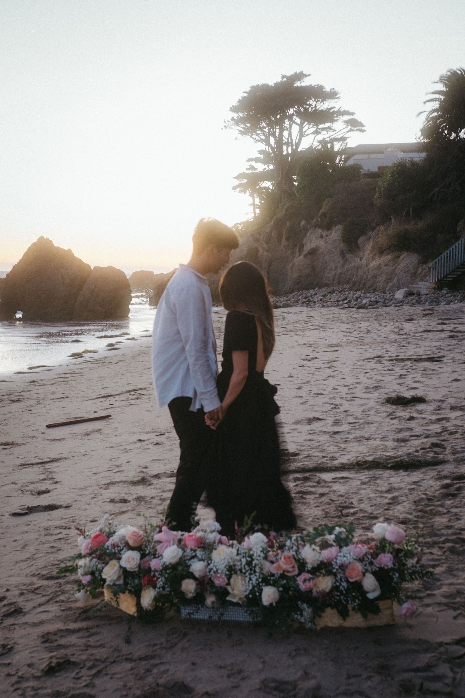 Romantic couple holding hands at sunset on beach with beautiful floral arrangement in foreground.