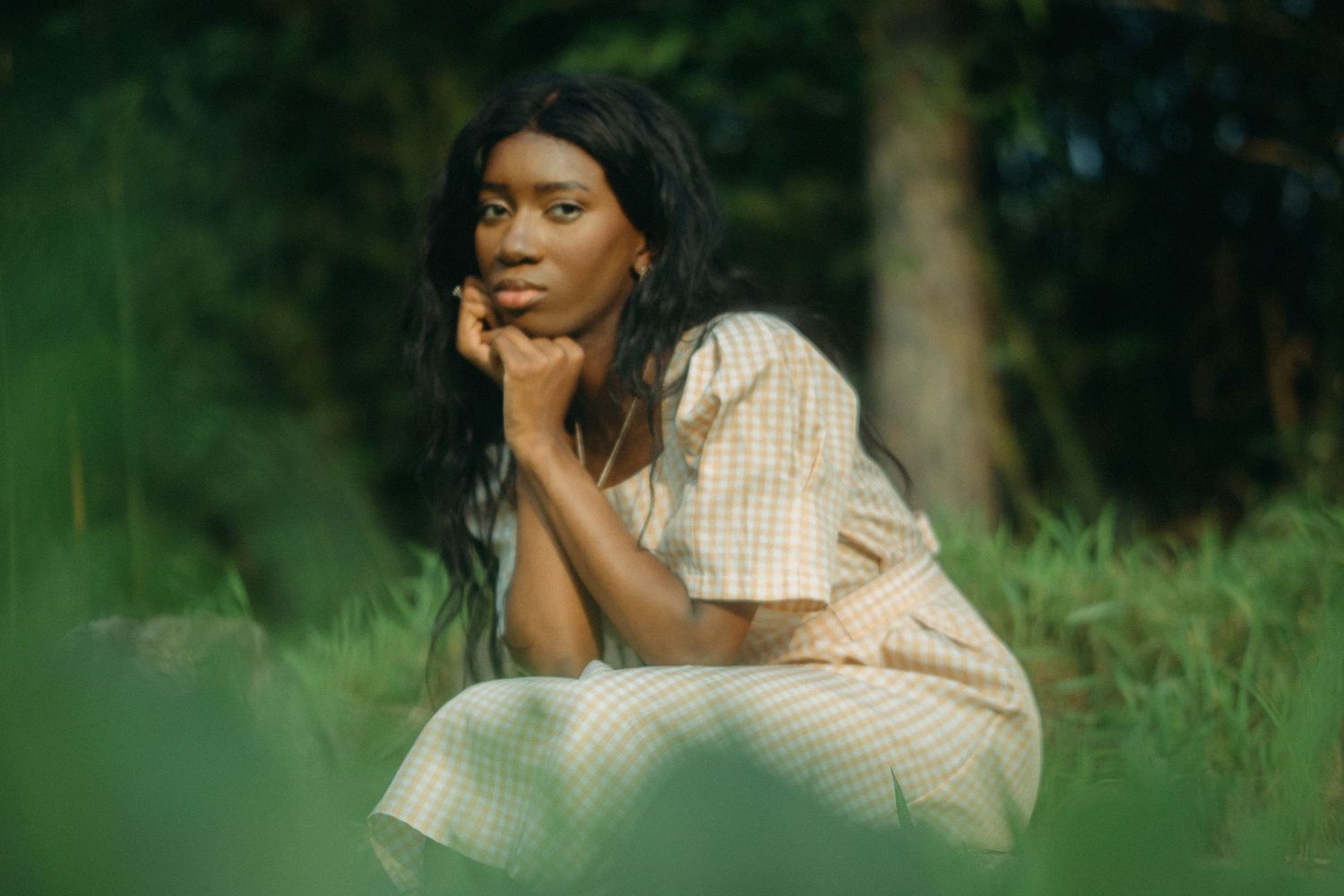 Thoughtful young woman in checkered dress sitting in green grass looking away.