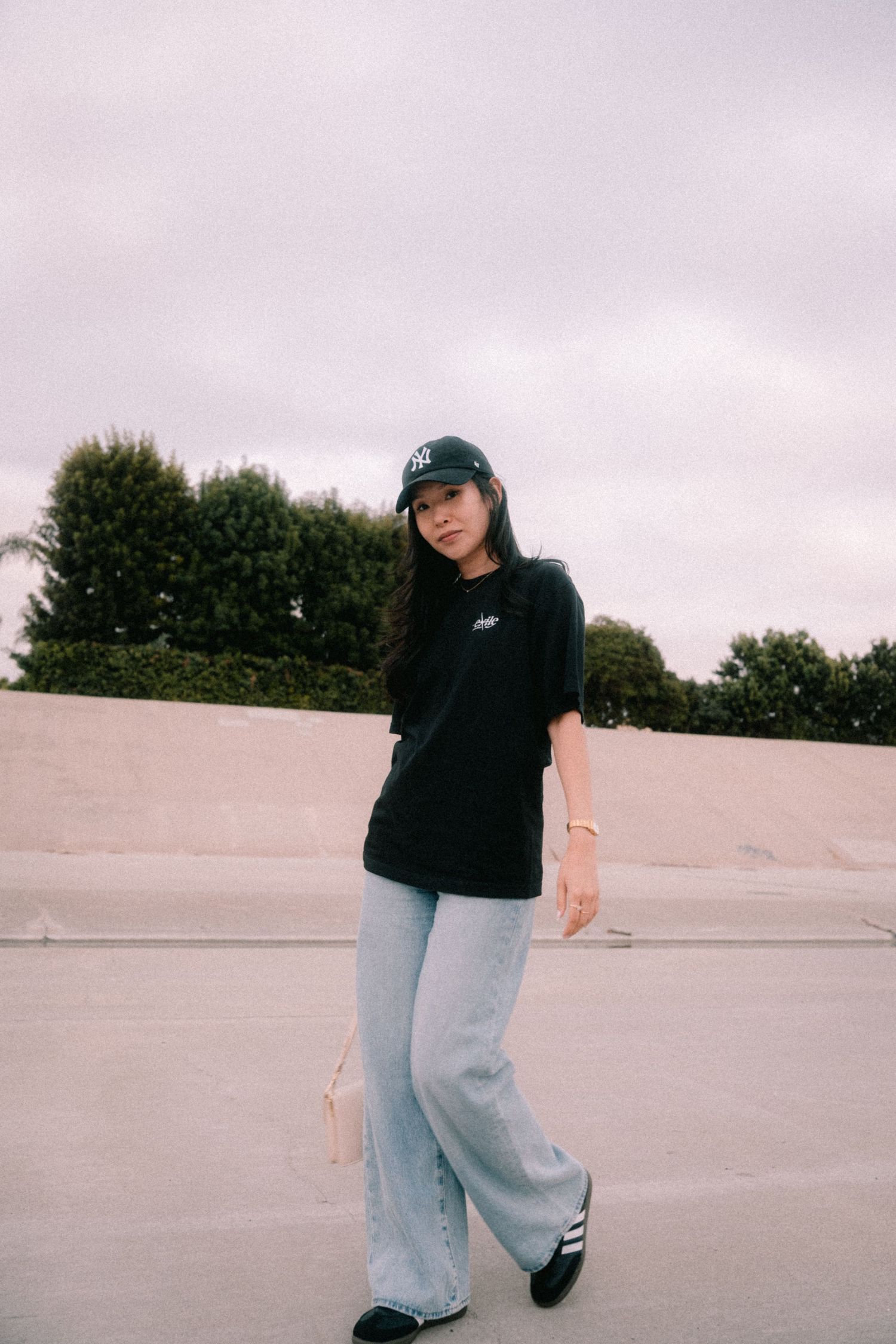 Woman in black oversized tee, wide-leg jeans, and baseball cap posing at an outdoor skate park.