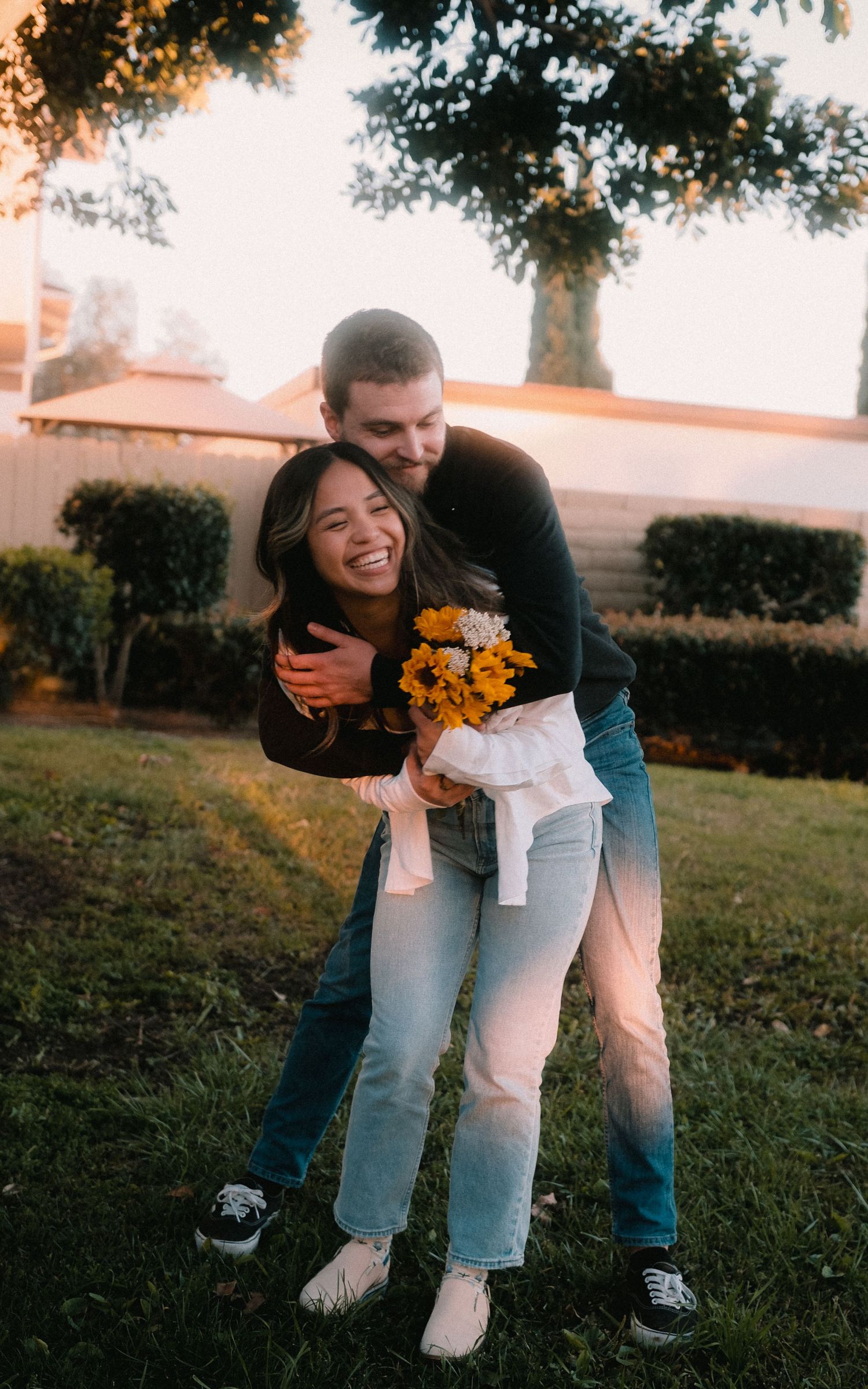 A joyful couple embraces in a park at sunset, woman laughing while holding yellow flowers.