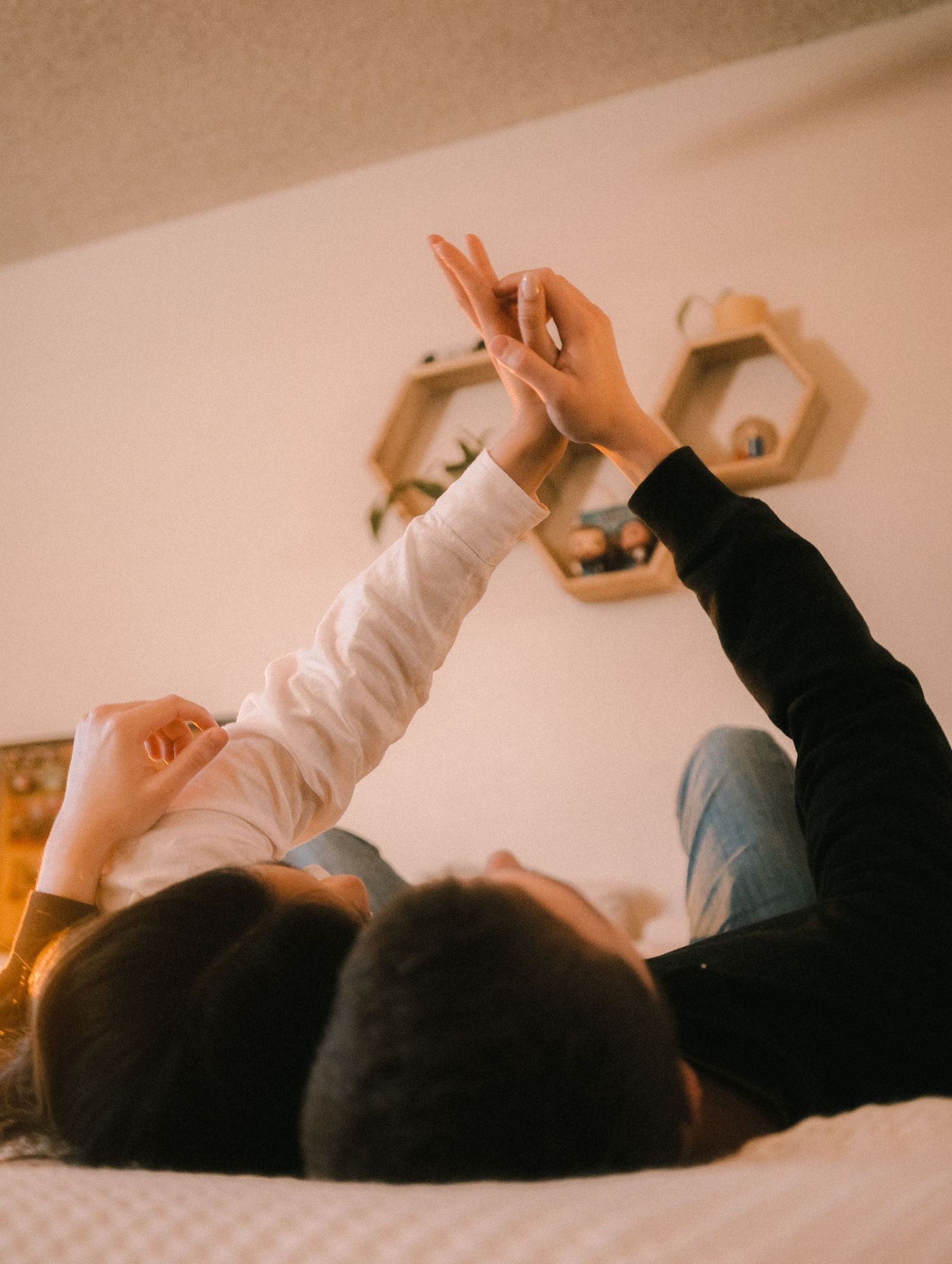 Couple lying on bed holding hands up toward ceiling with hexagonal shelves on wall behind them.