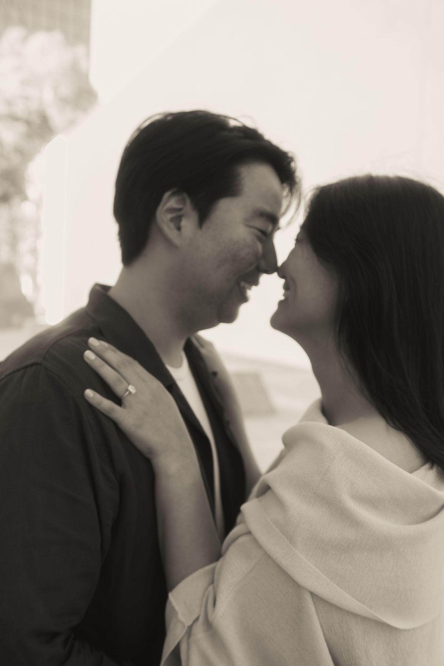 Black and white photo of an Asian couple nearly kissing, woman displaying engagement ring.