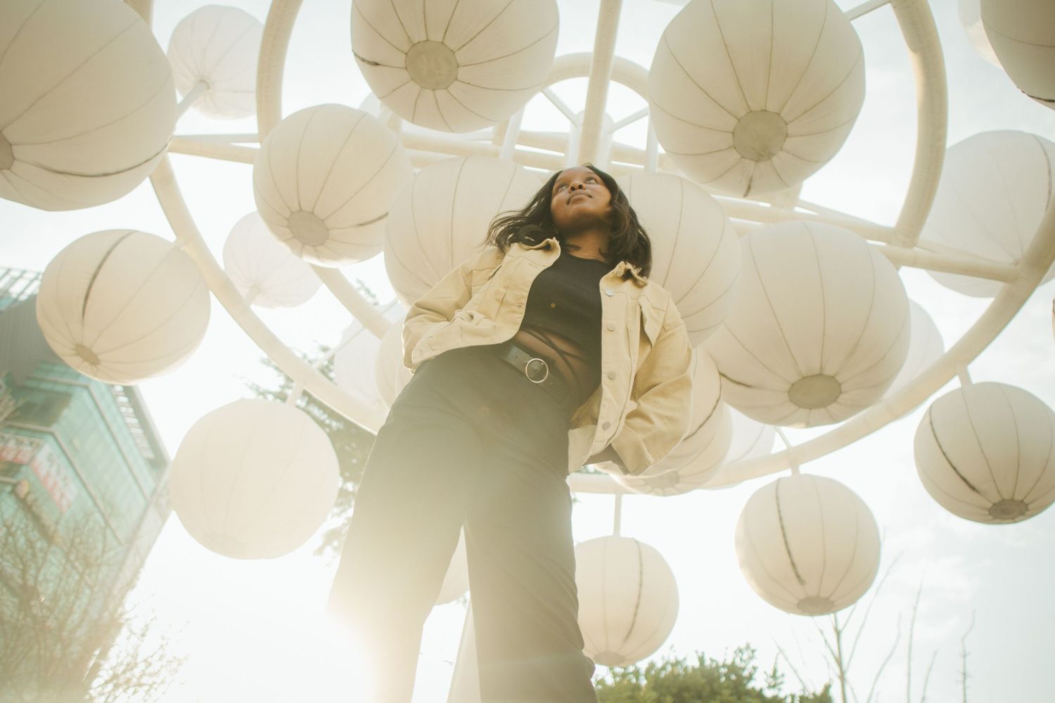 Woman standing among white paper lanterns in an outdoor art installation, shot from below against bright sky.