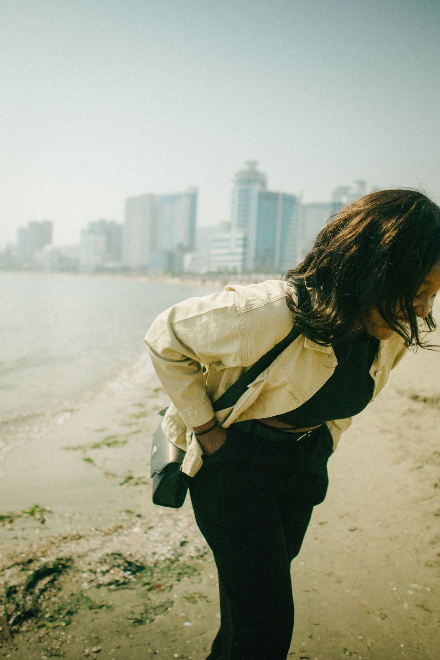 Woman in yellow jacket bending forward on a beach with a city skyline in the background.