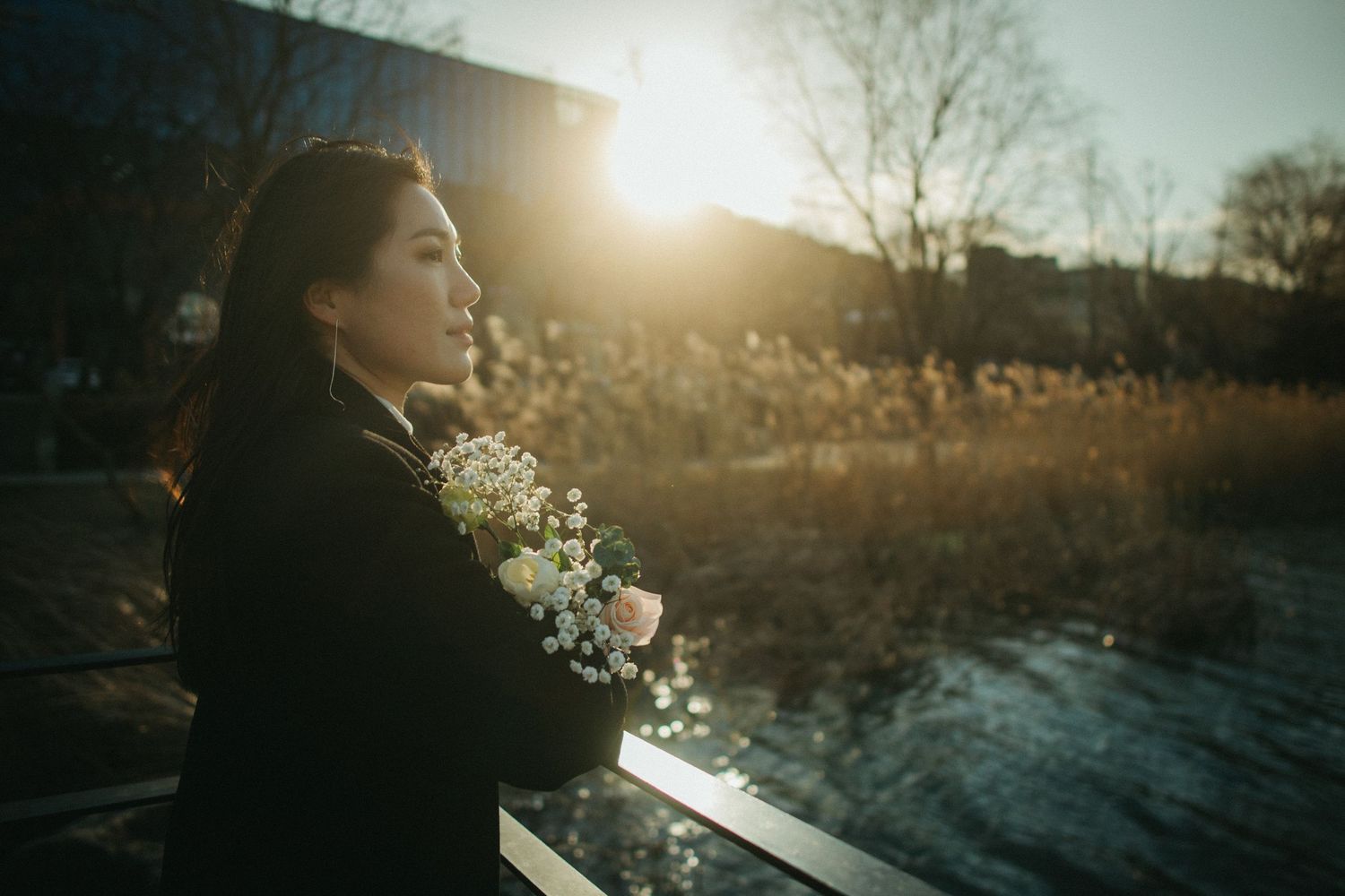 Young woman in black coat holding white flowers backlit by golden sunset light outdoors.
