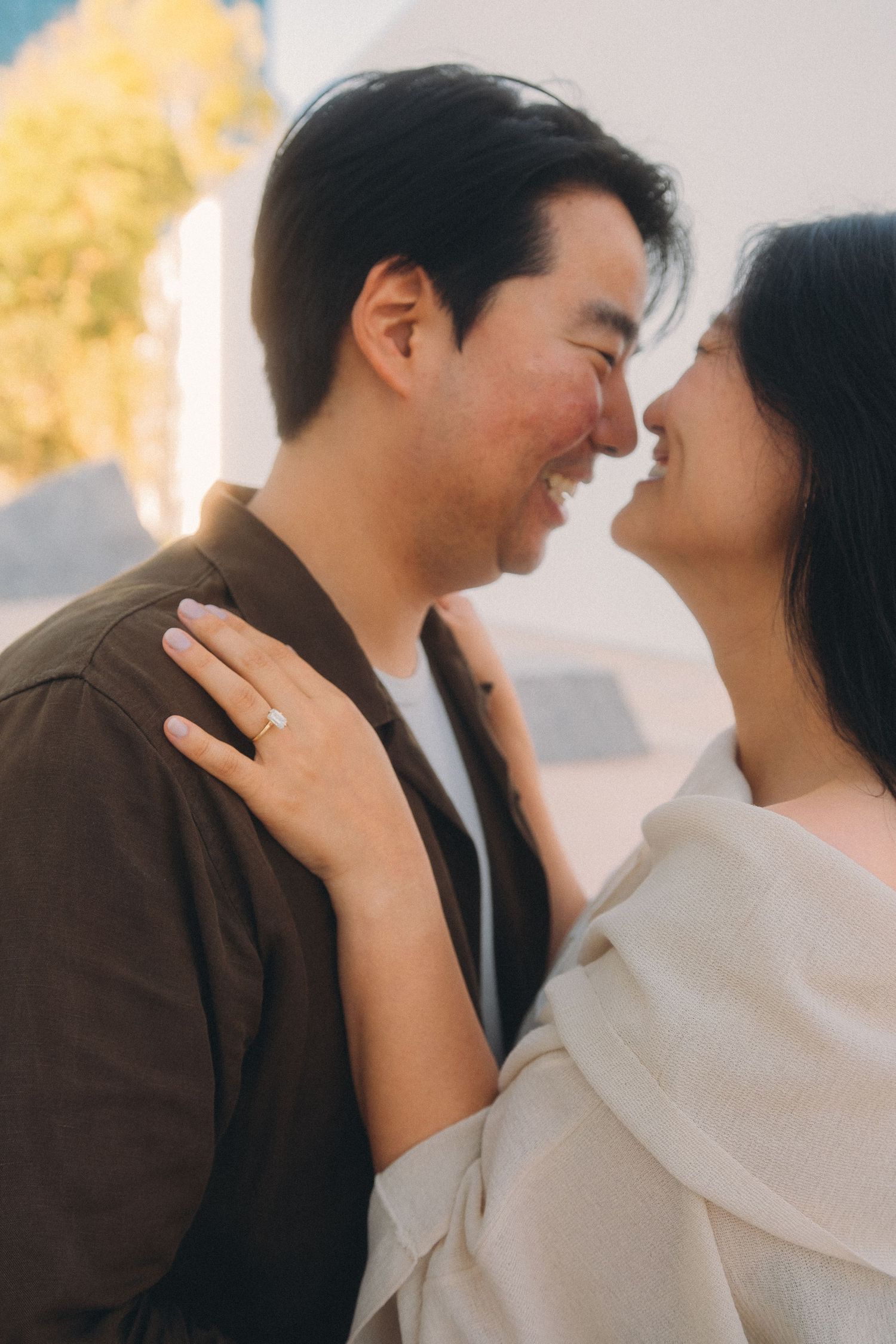 A smiling couple shares a romantic moment, the woman wearing an engagement ring, bathed in warm golden sunlight.