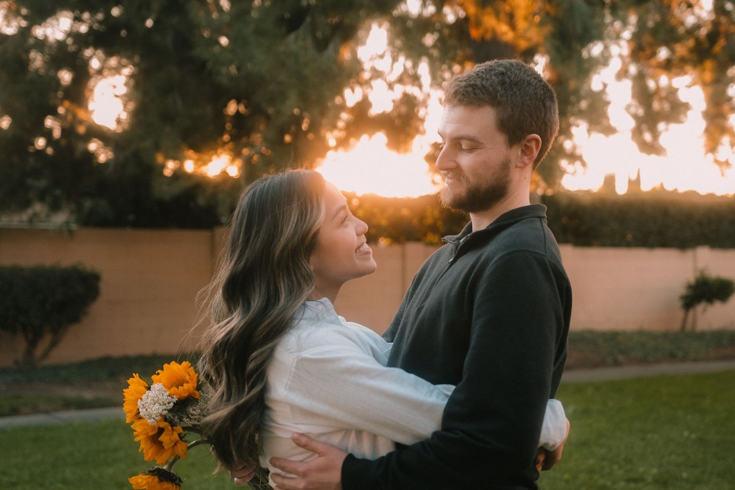 Couple embracing at sunset in a garden, woman holding sunflowers, golden backlight through trees.