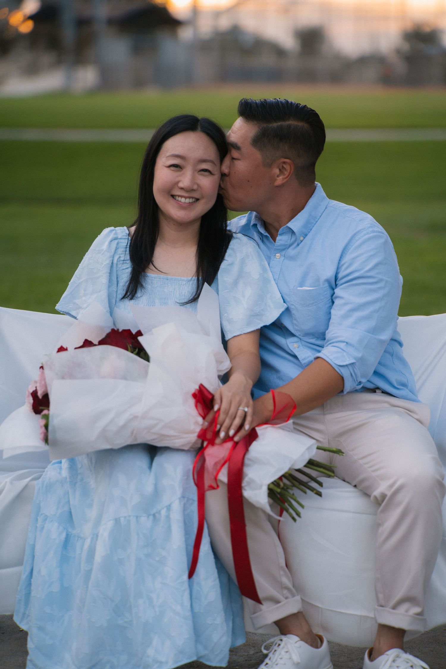 Couple sitting on white couch outdoors, man kissing woman's cheek as she smiles holding red roses bouquet.
