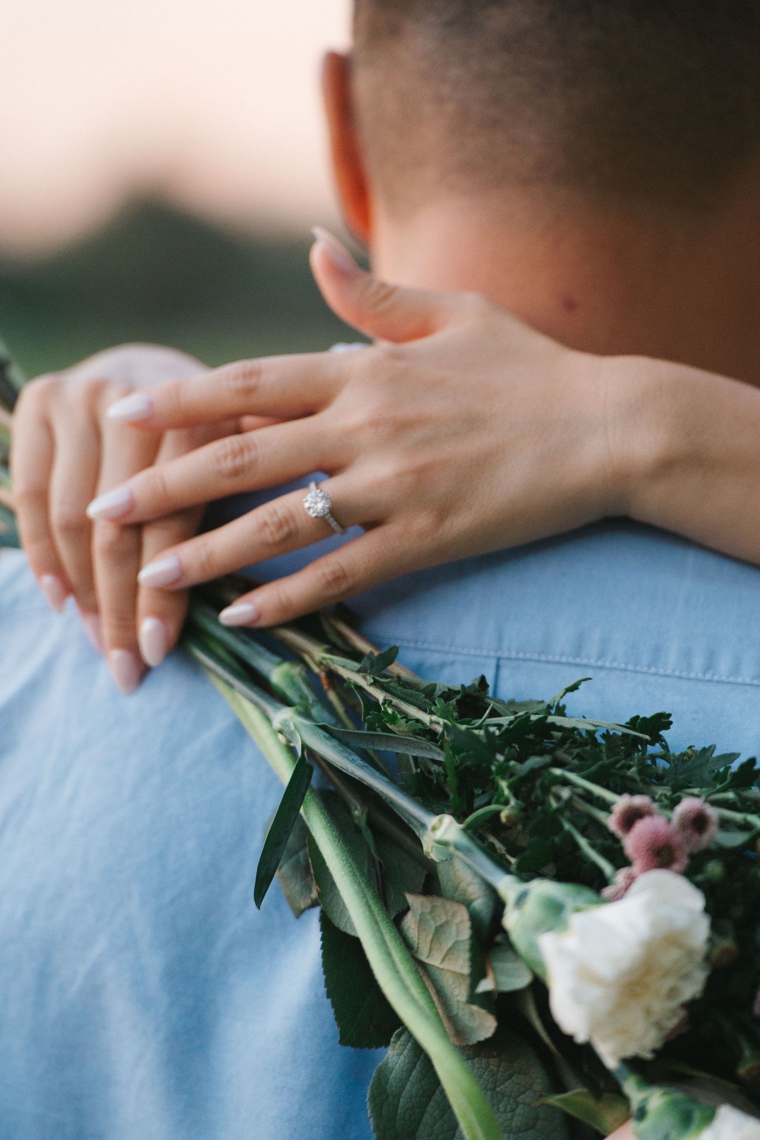 Woman wearing diamond engagement ring embracing man while holding a bouquet of white flowers and greenery.