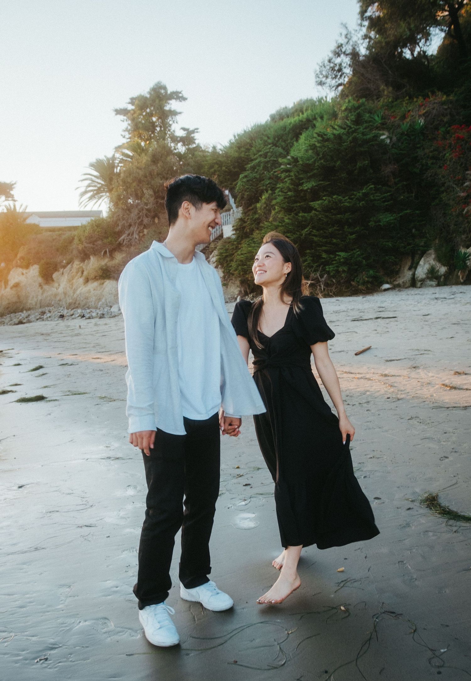 Happy couple holding hands and smiling at each other while walking on a sandy beach at sunset.