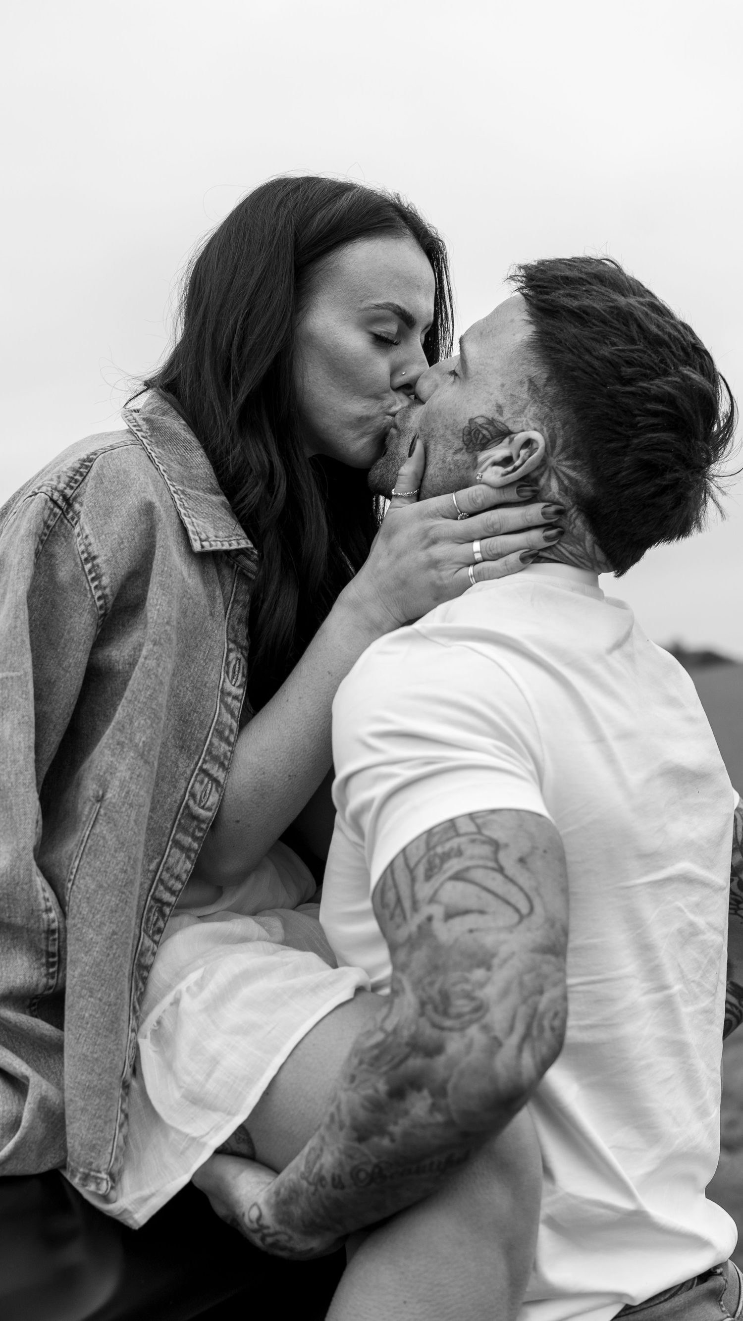 Black and white photo of a tattooed couple sharing a passionate kiss outdoors in a field setting.