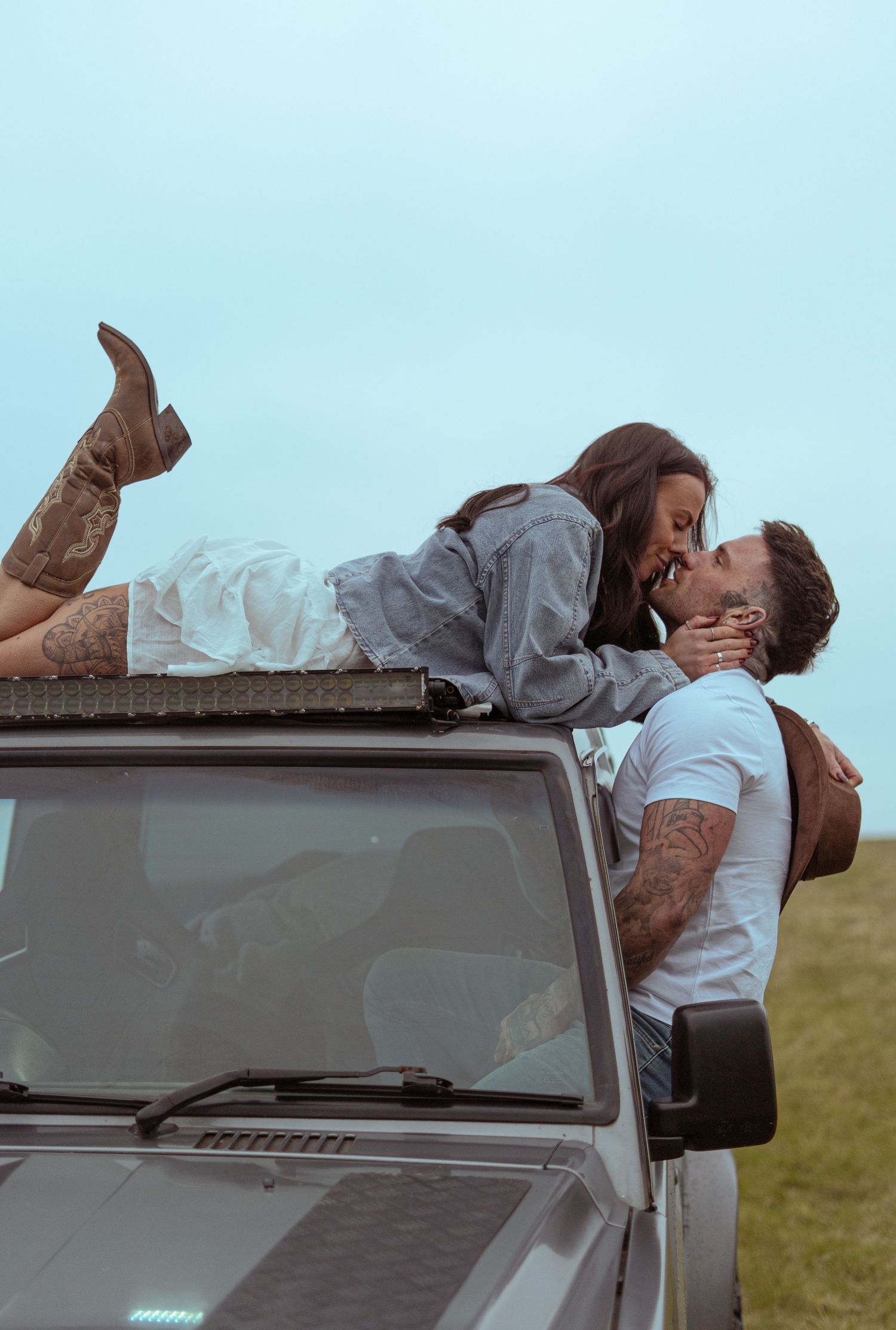 Couple sharing a romantic kiss on the hood of a vintage off-road vehicle in an open field setting.