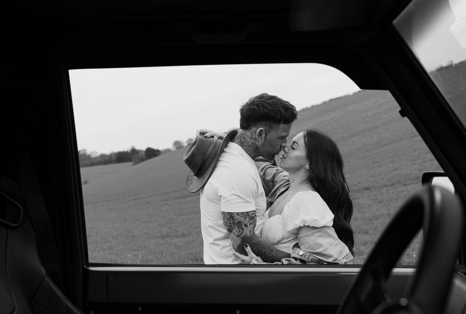 Black and white photo of a couple kissing outside, framed through a vintage vehicle window in a field.
