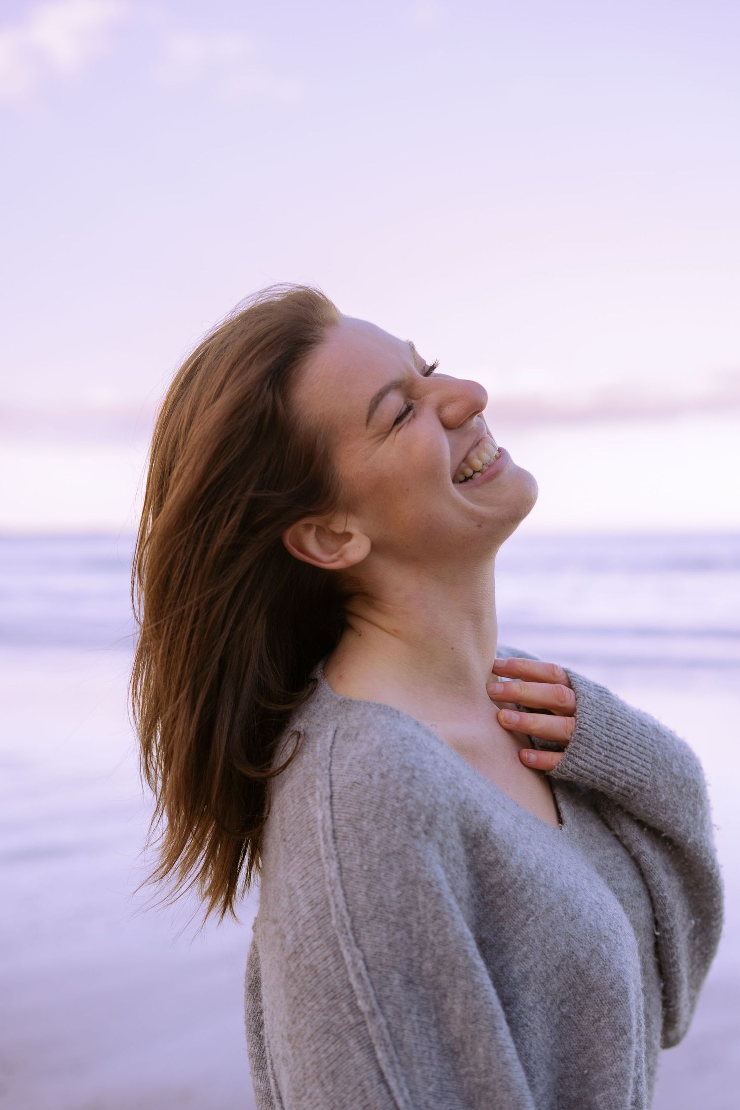 Happy woman laughing with eyes closed on a beach at dusk in a grey sweater.