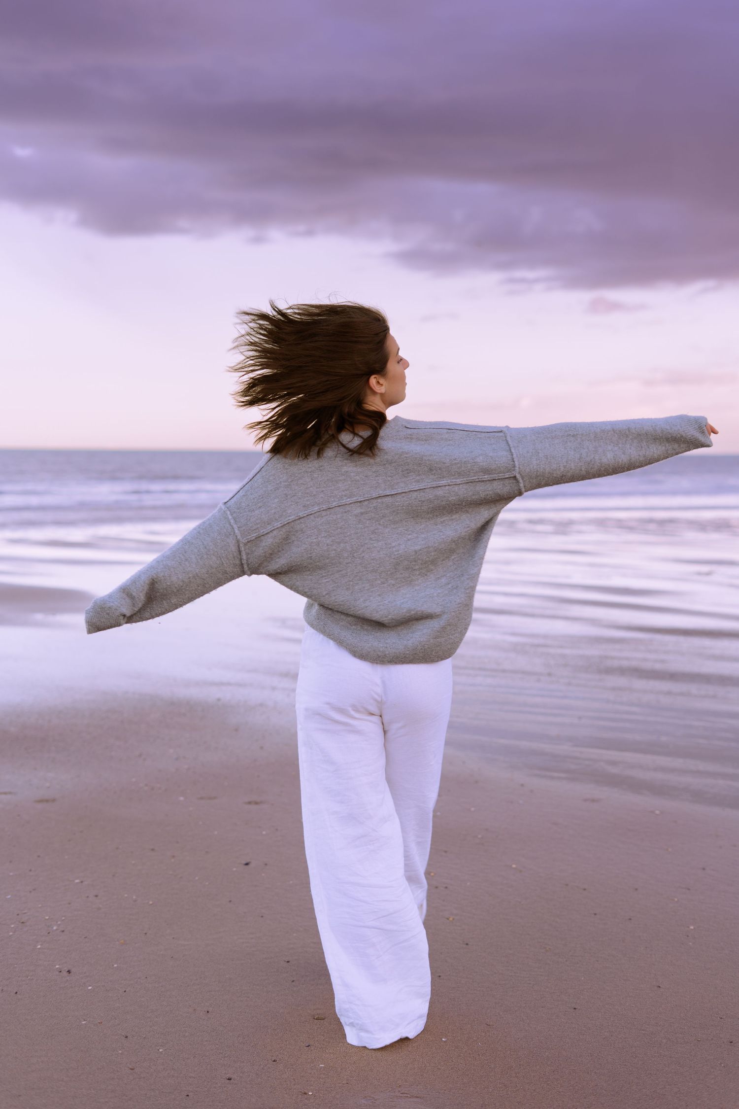 Free-spirited woman in grey sweater and white pants standing arms outstretched on a purple sunset beach.