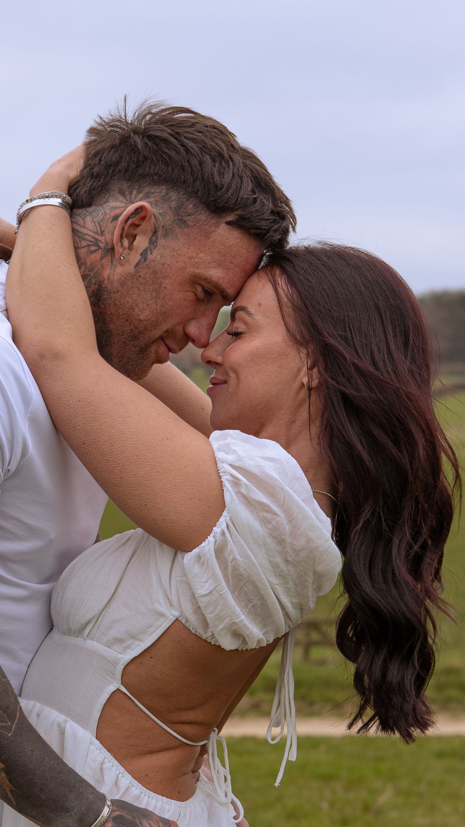 Couple sharing a tender forehead-to-forehead embrace outdoors, woman in white top with long dark hair.