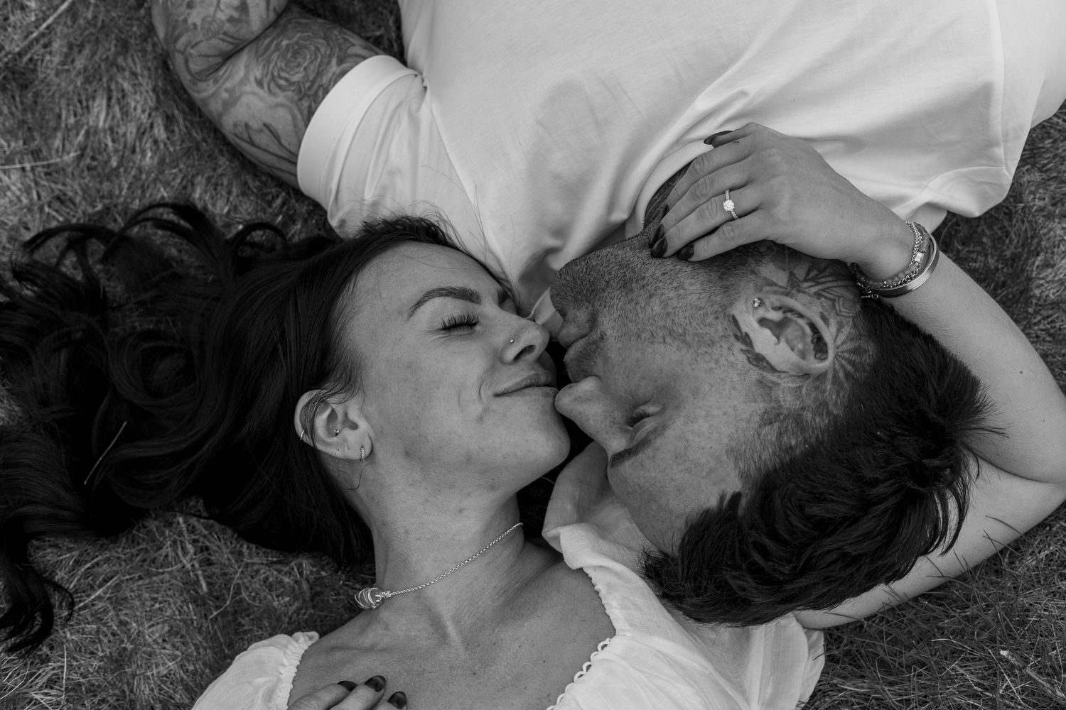 Black and white photo of a couple lying in grass, sharing a tender almost-kiss in a romantic outdoor moment.